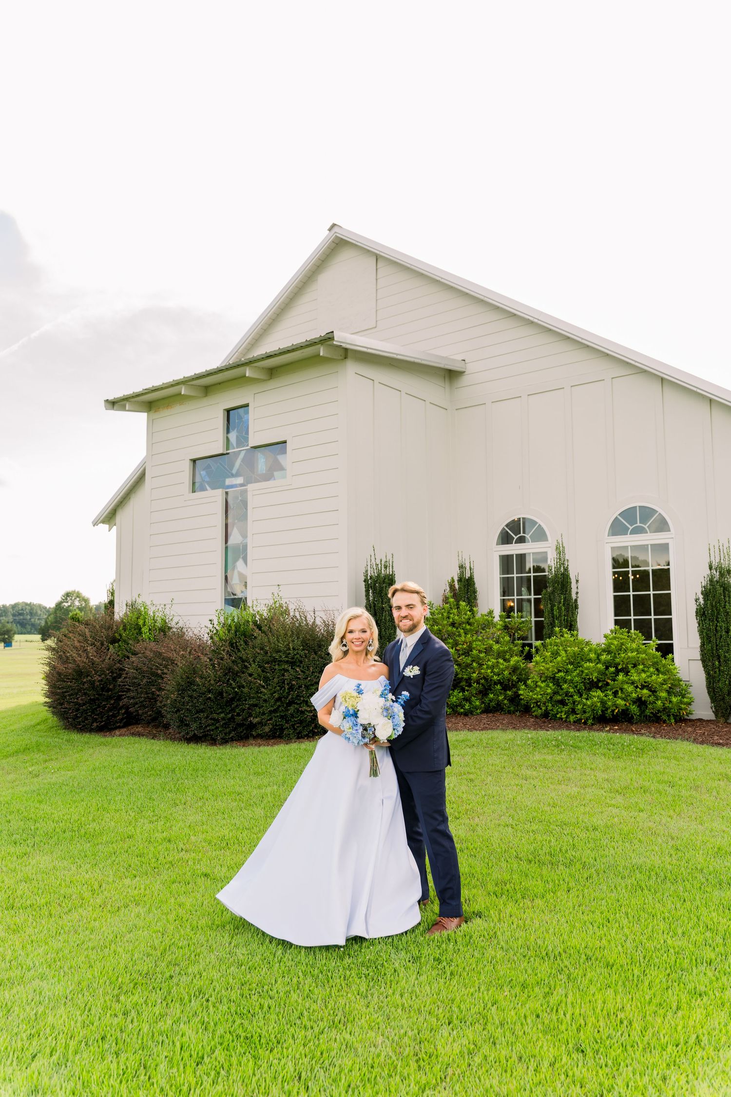 Bride and groom pose together on green lawn before a white chapel with a cross on their wedding day.