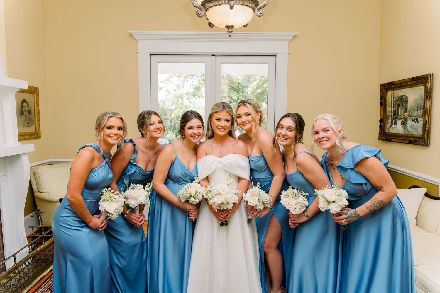 Bride in white gown poses with six bridesmaids in blue satin dresses holding white bouquets indoors.