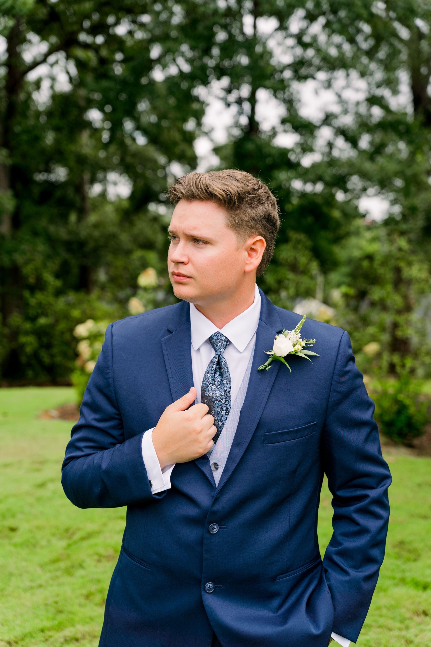 Groom in navy blue suit with floral boutonniere and patterned tie poses outdoors in lush green garden setting.