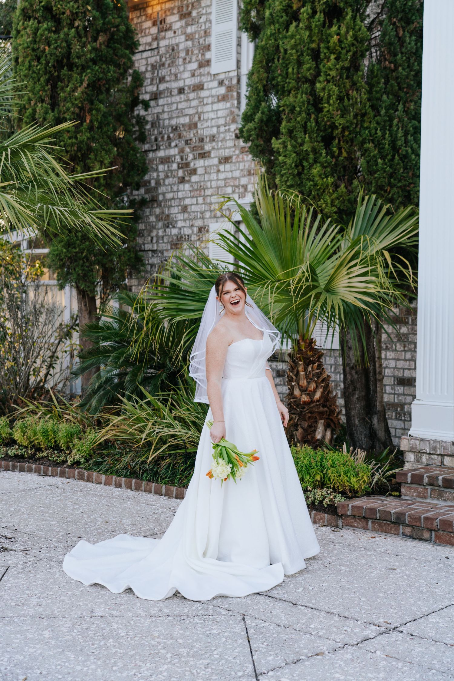 A flowing white wedding dress is photographed against a brick wall backdrop with lush greenery.