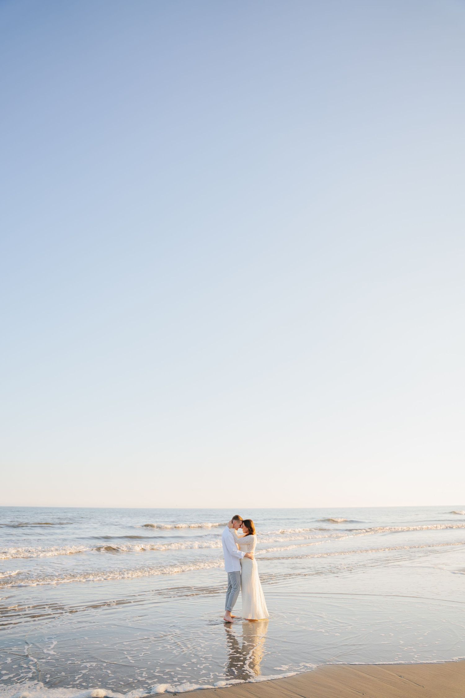 Person in white clothing walking along a serene beach shoreline at sunset with waves gently rolling in.