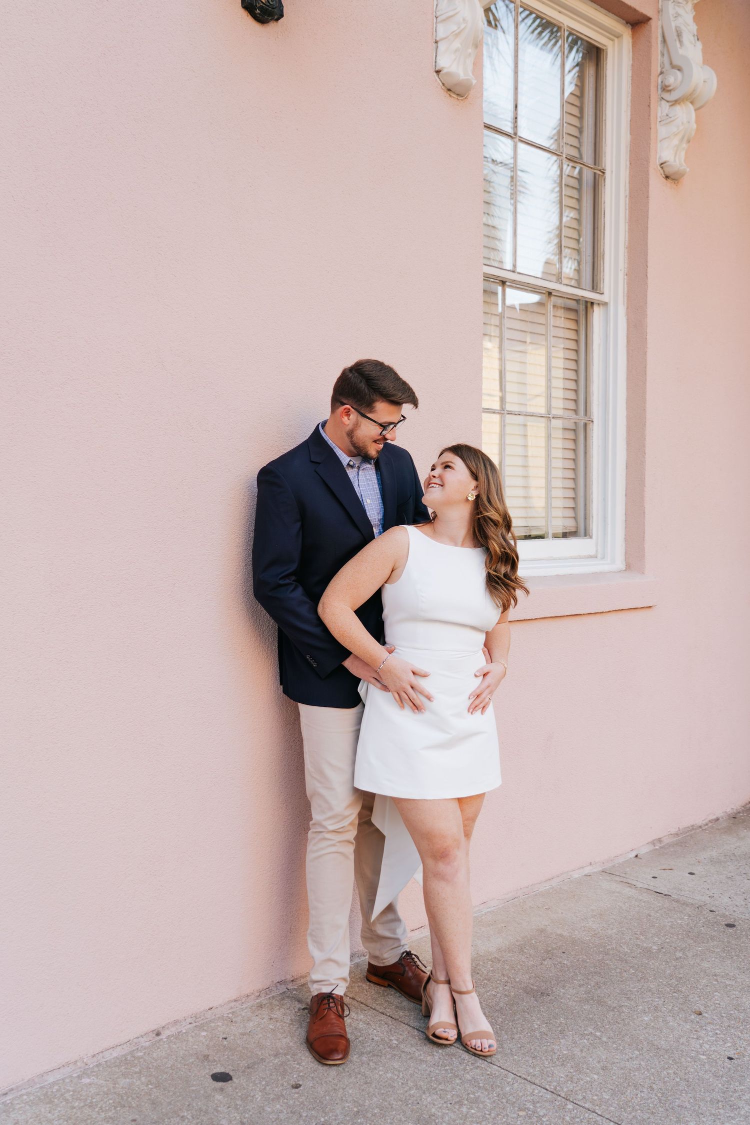 A romantic couple poses against a pink wall wearing coordinated white and navy outfits.