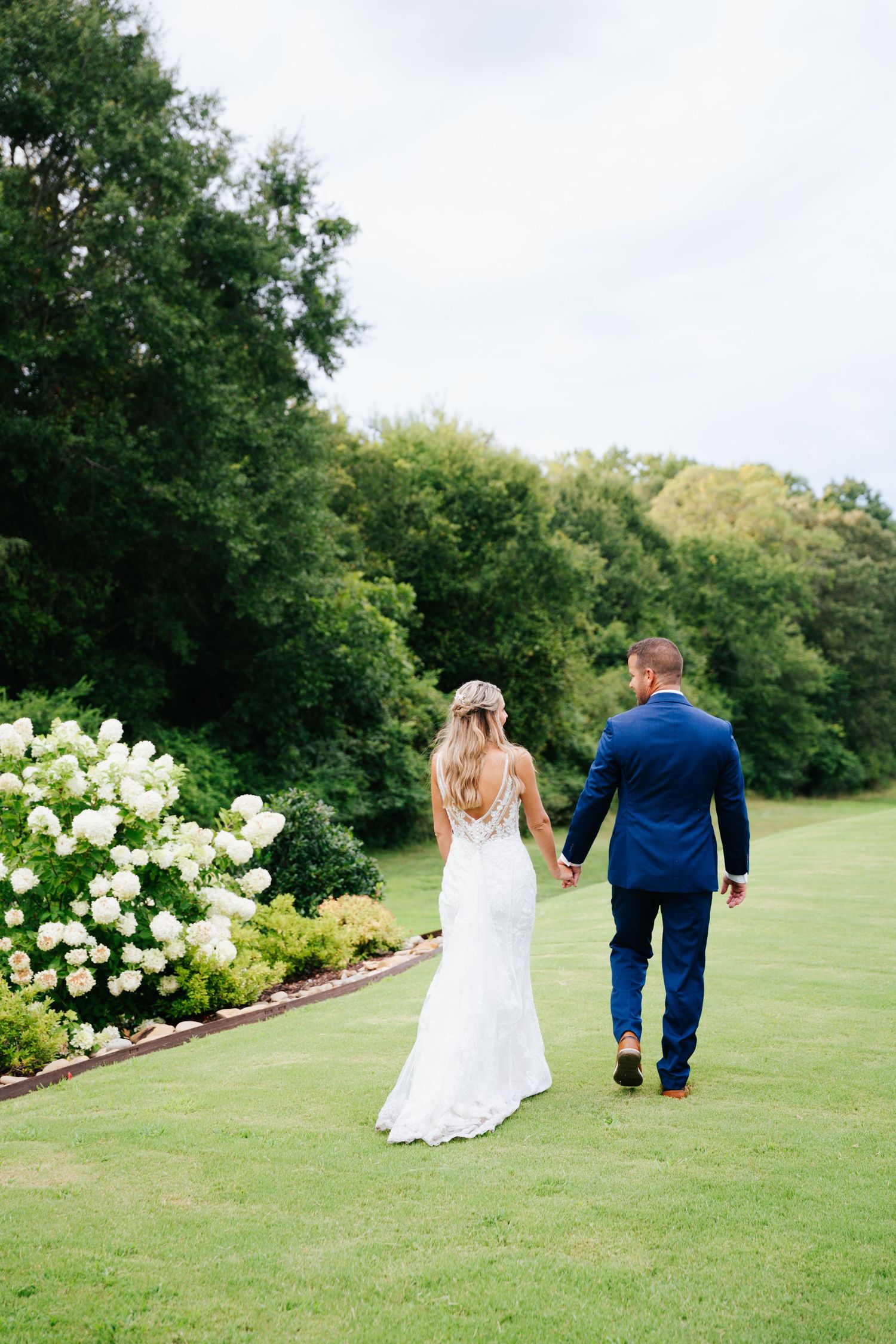 Couple walks hand in hand across a lush green lawn with white flowers in background.