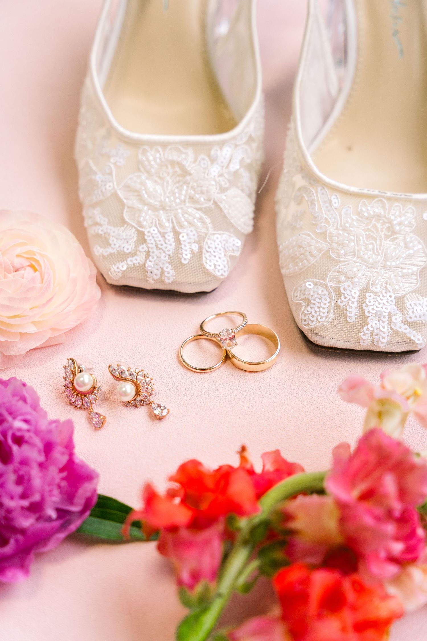 White lace bridal heels with gold wedding rings, pearl earrings, and colorful flowers on a pink background.