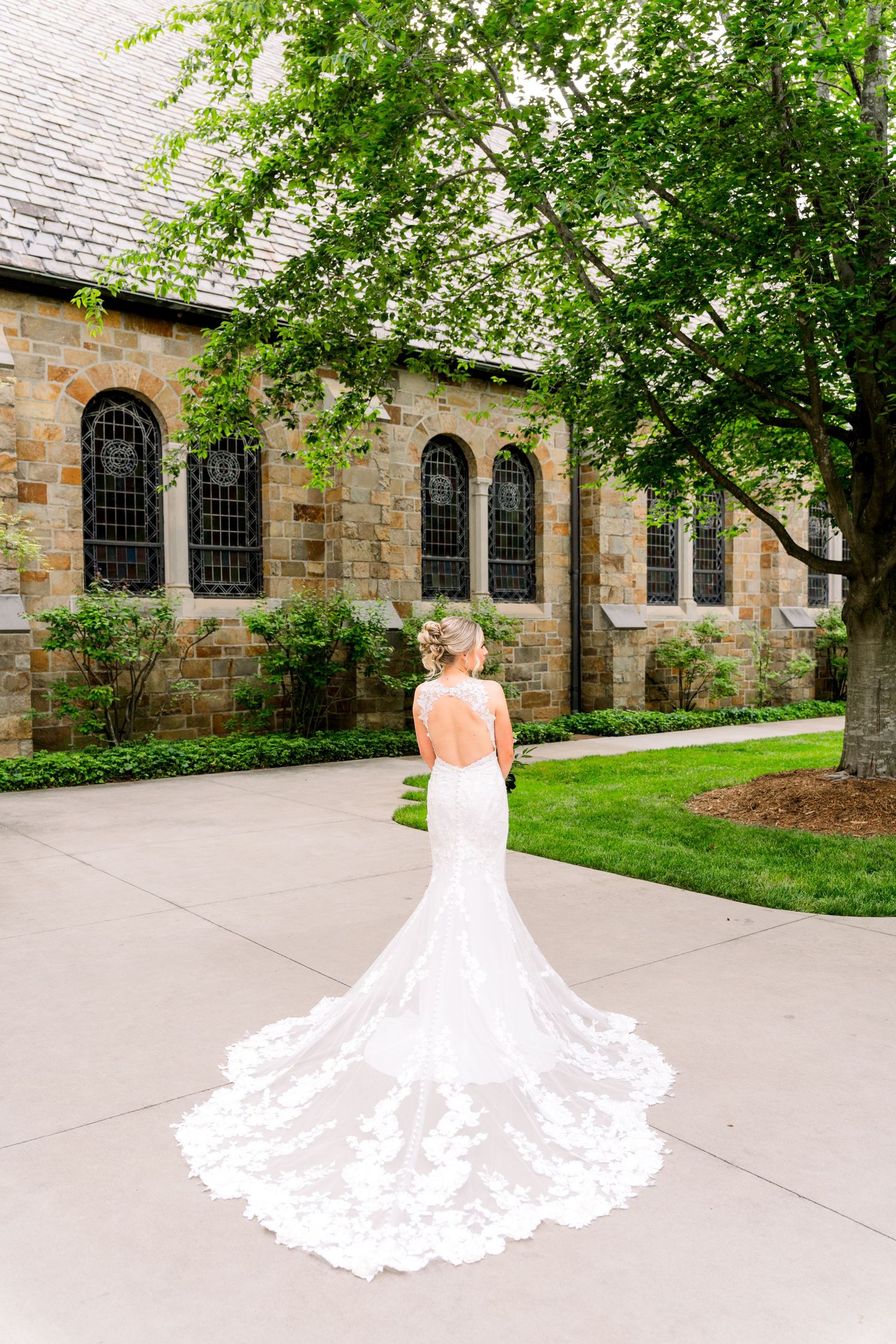 Bride in backless lace wedding gown with cathedral train stands before stone church surrounded by lush green trees.