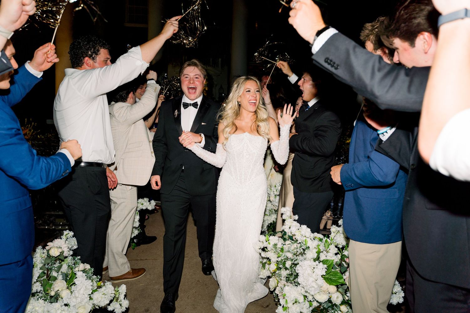 Bride and groom exit wedding reception through sparkler send-off surrounded by cheering guests in formal attire.