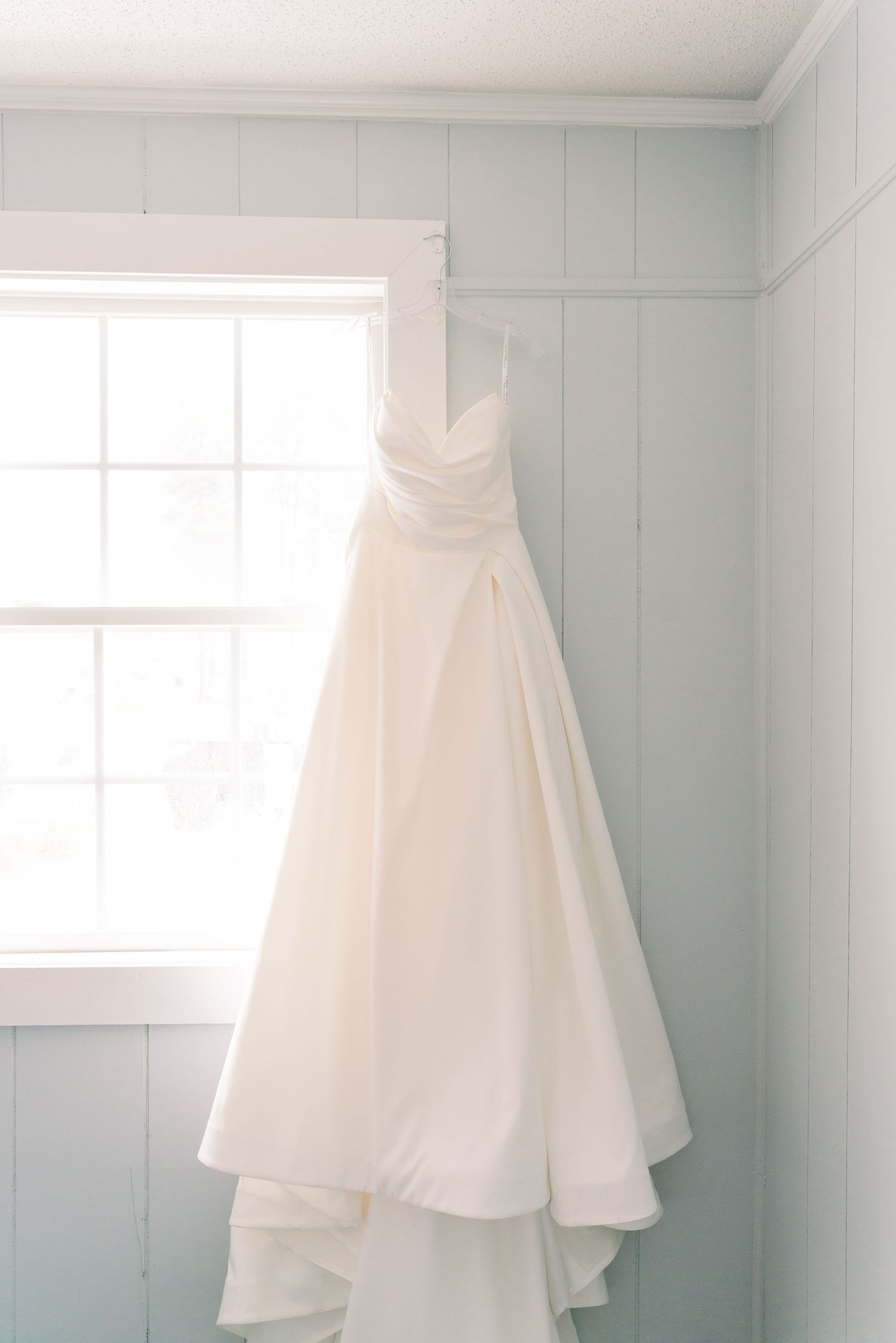 A white wedding dress hangs elegantly against a pale blue wall next to a bright window, showcasing its flowing train.