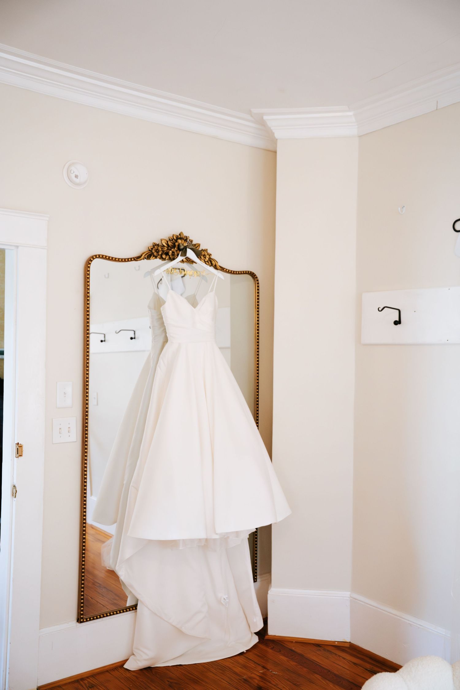 A white wedding dress and smaller dress hang on display against cream colored walls in a room with hardwood floors.