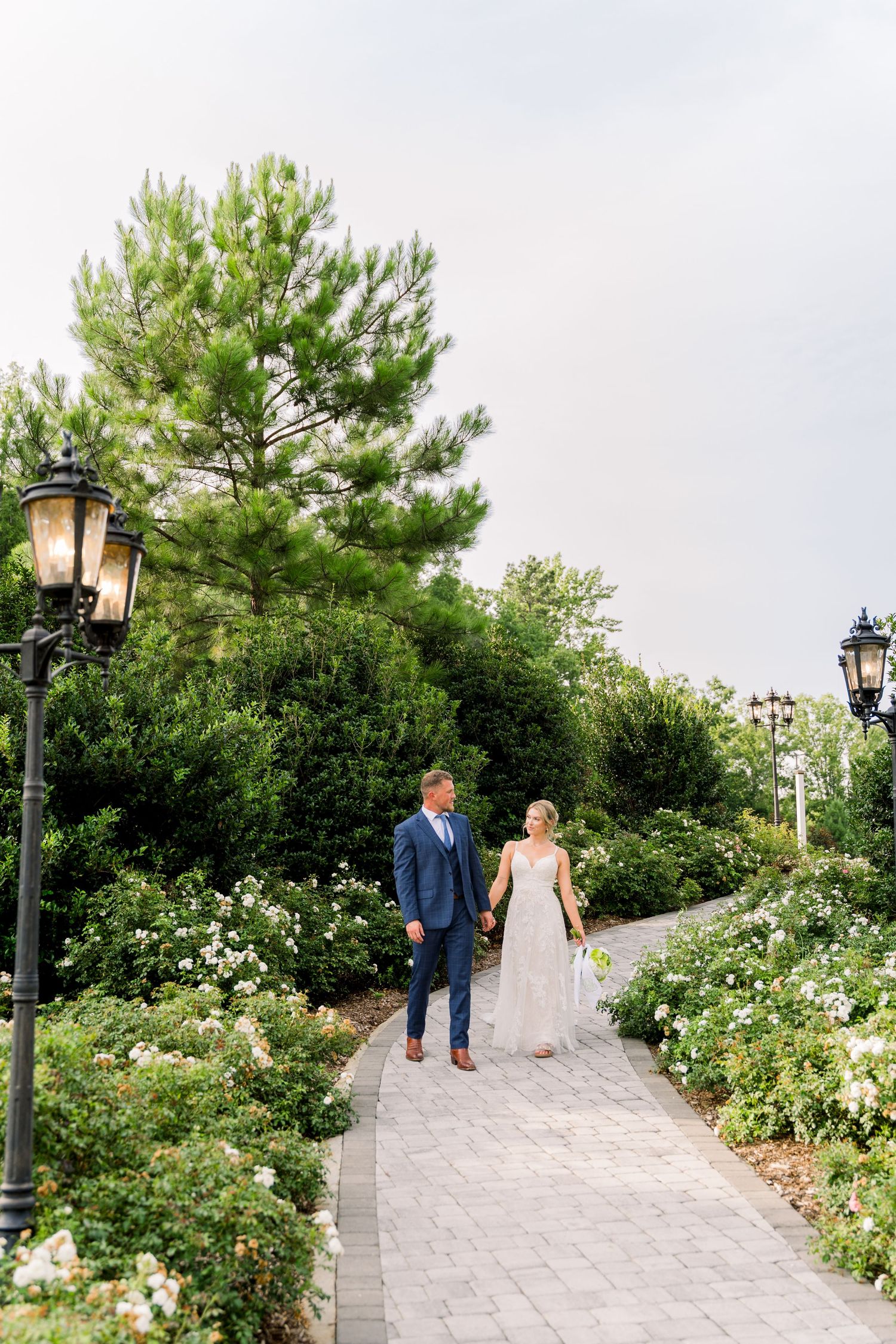 Bride and groom walk hand-in-hand along a garden path lined with white roses and vintage lamp posts.