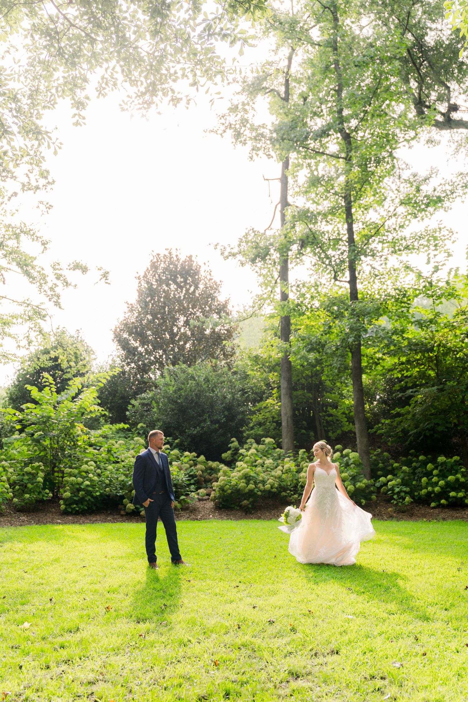 Bride in flowing white gown and groom in navy suit share a romantic moment on lush green lawn surrounded by trees.