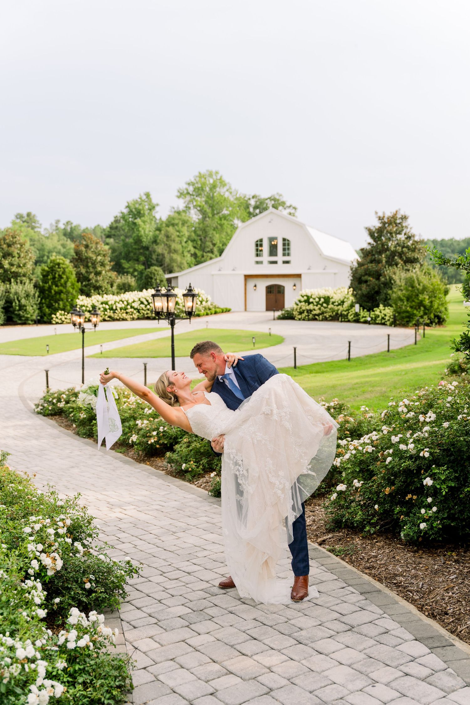 Groom dips bride for a kiss on a brick path surrounded by white flowers, with an elegant white barn venue in the background.