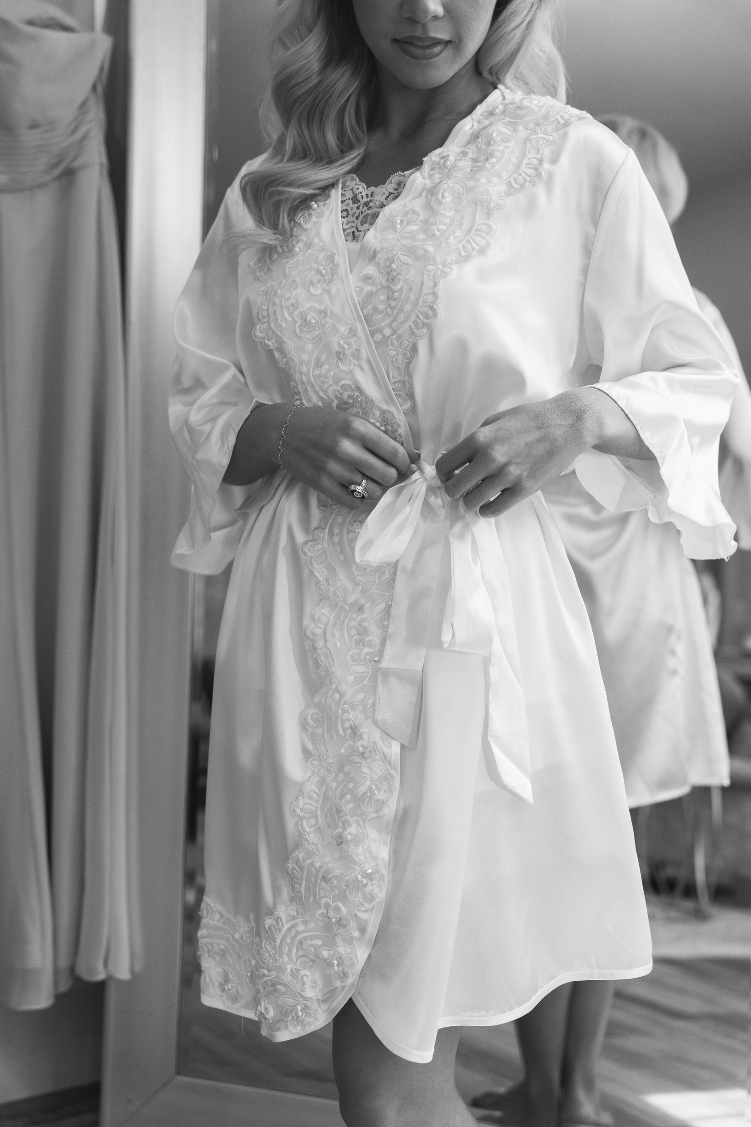 Black and white photo of bride in lace-trimmed white satin robe adjusting her belt before the wedding.