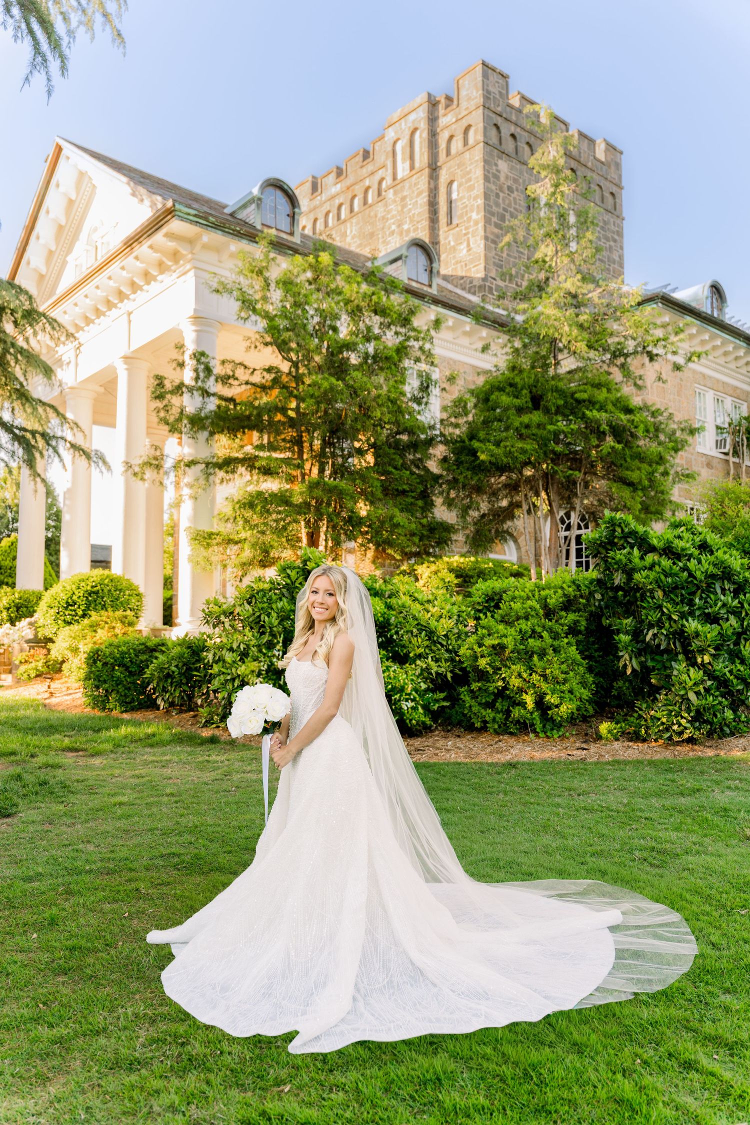 Bride in a flowing white gown and long veil holds bouquet on green lawn before elegant mansion.