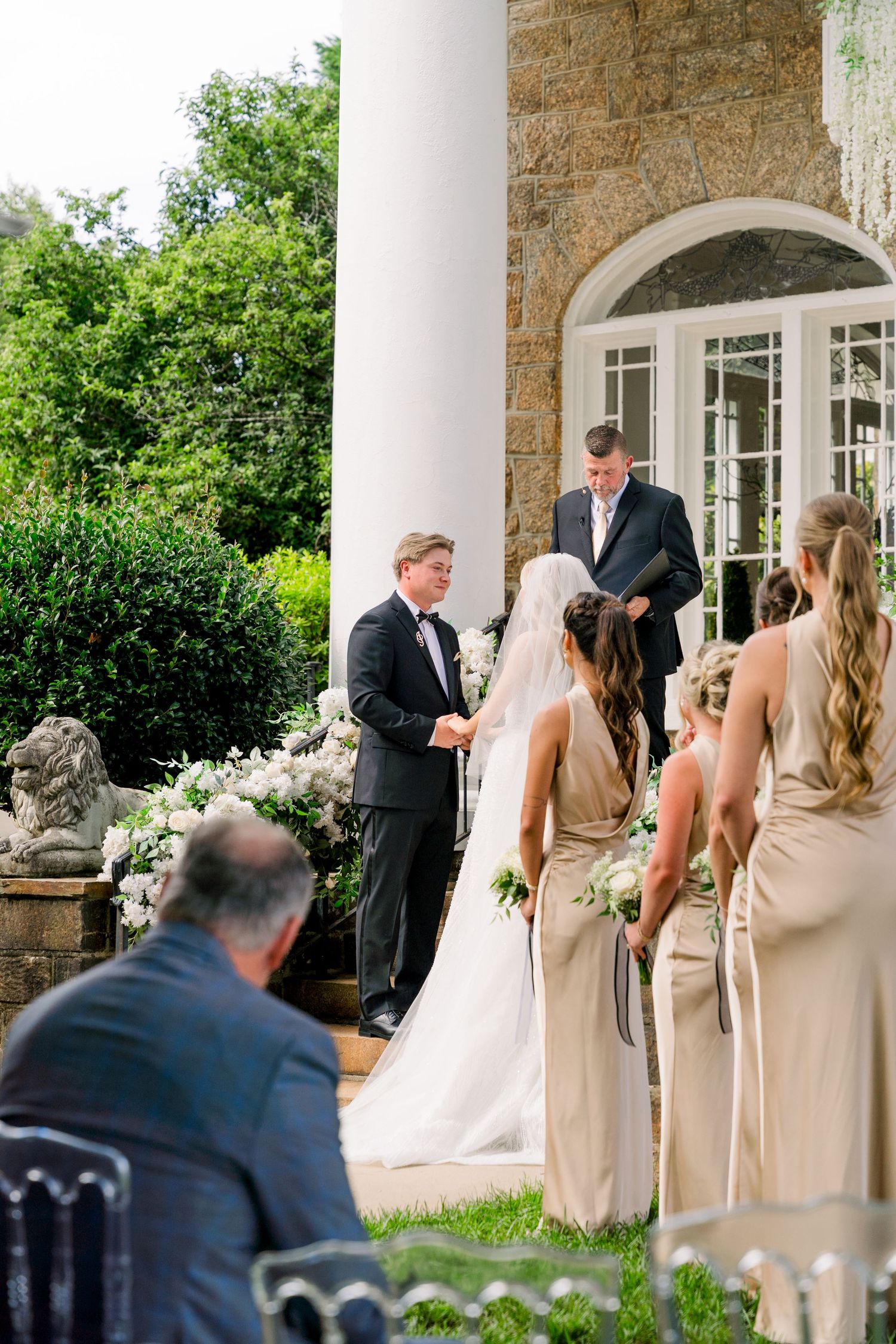 Outdoor wedding ceremony with bride in white gown, groom in tuxedo, bridesmaids in champagne dresses, officiant reading vows.
