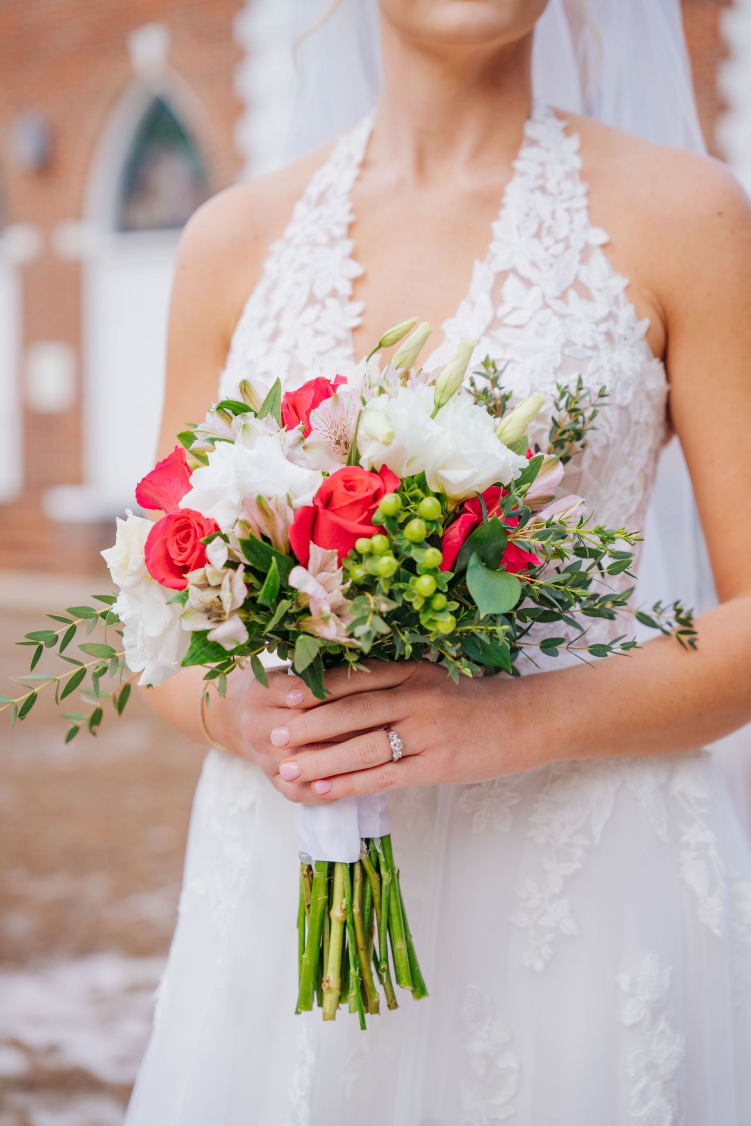 A bridal bouquet featuring pink and white roses with greenery is held against a white lace wedding dress.