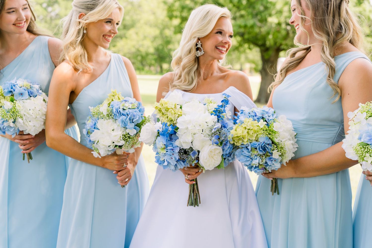 Bride in white gown with bridesmaids in light blue dresses holding blue and white hydrangea bouquets outdoors.