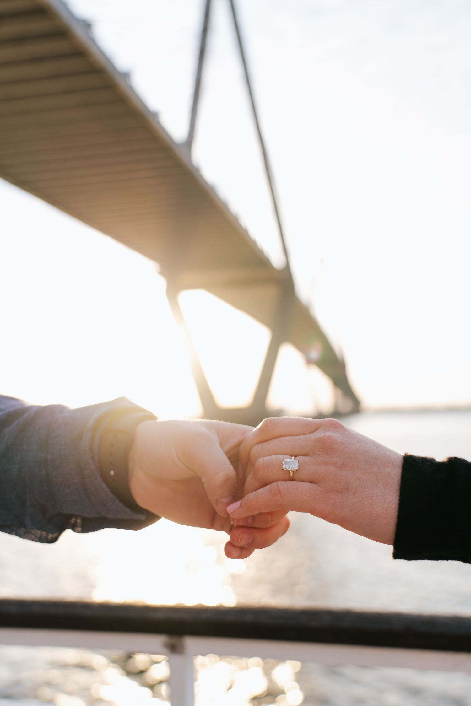 Close-up of hands displaying an engagement ring with a bridge silhouetted in the background.