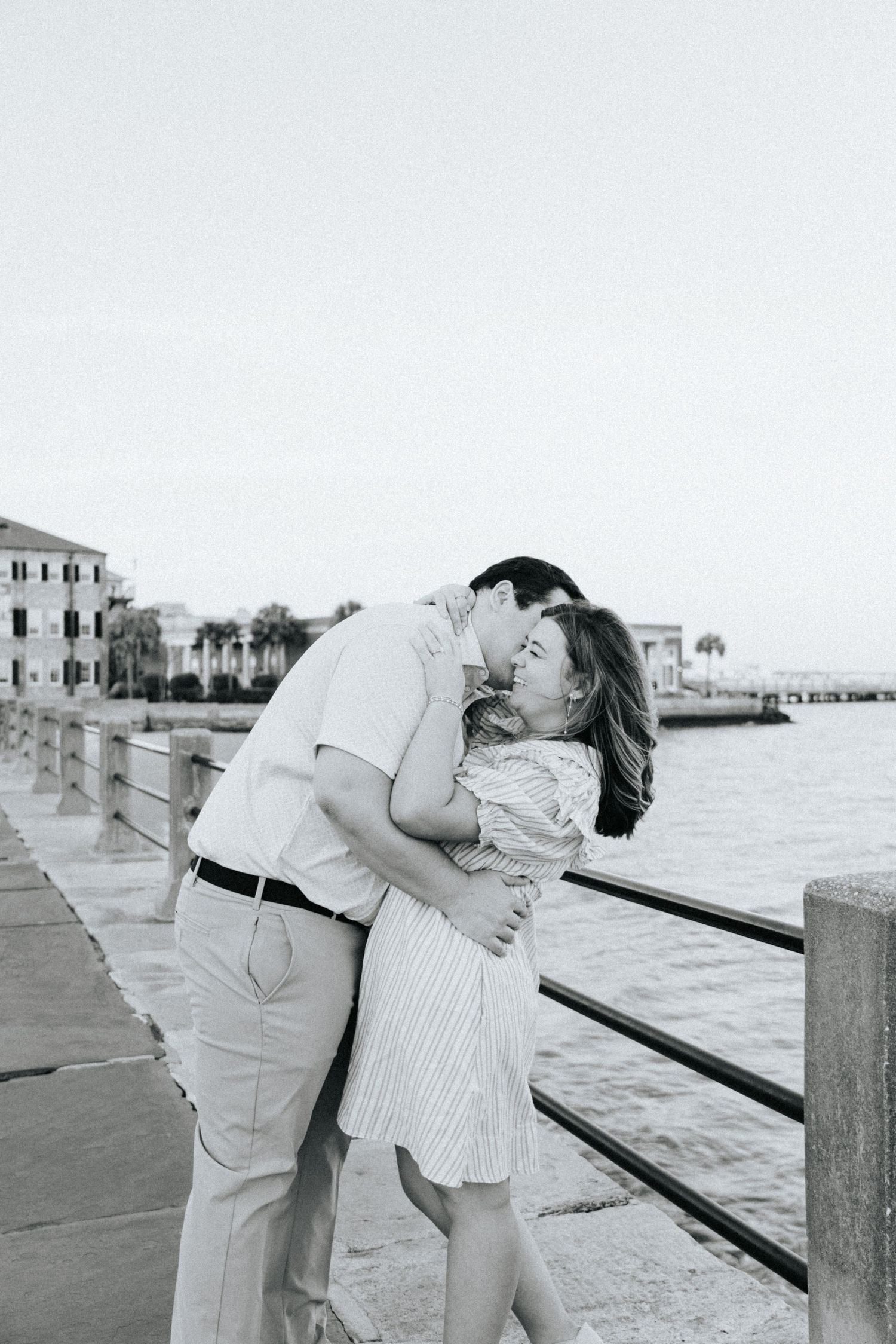 Black and white photo of couple sharing intimate moment along waterfront railing.