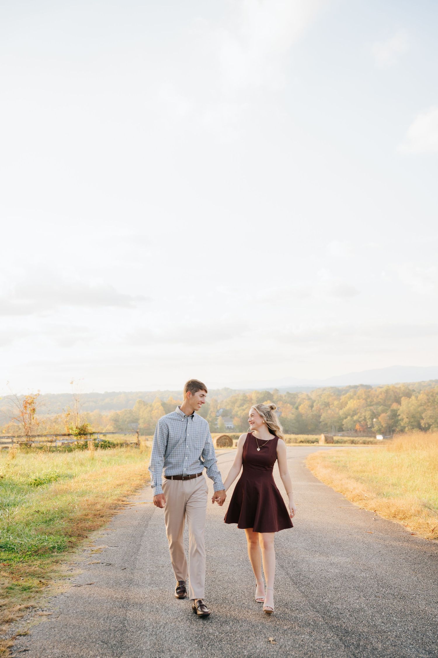 A couple walks hand in hand down a dirt path surrounded by autumn foliage and golden fields at sunset.