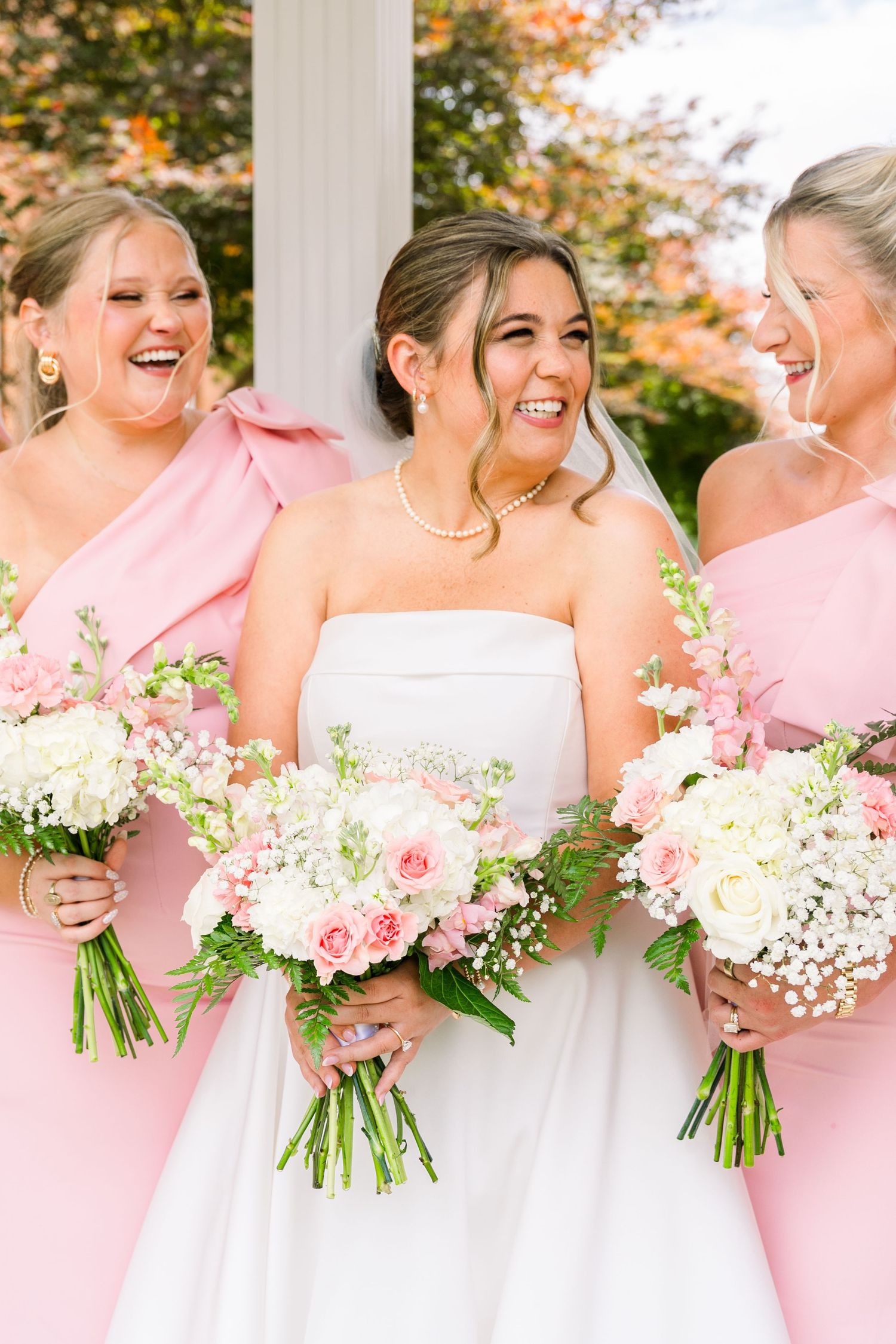 Bride in white strapless gown laughing with bridesmaids in pink dresses holding pink and white floral bouquets.