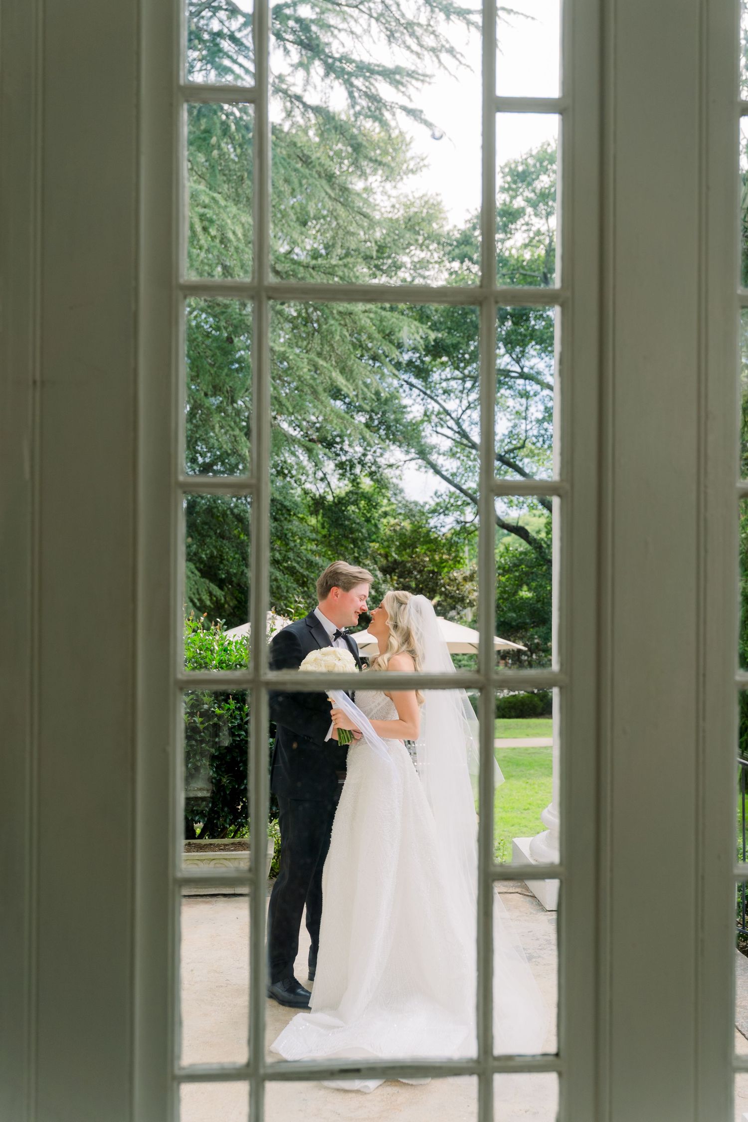 Bride and groom share a romantic kiss framed by elegant French doors overlooking a lush garden.