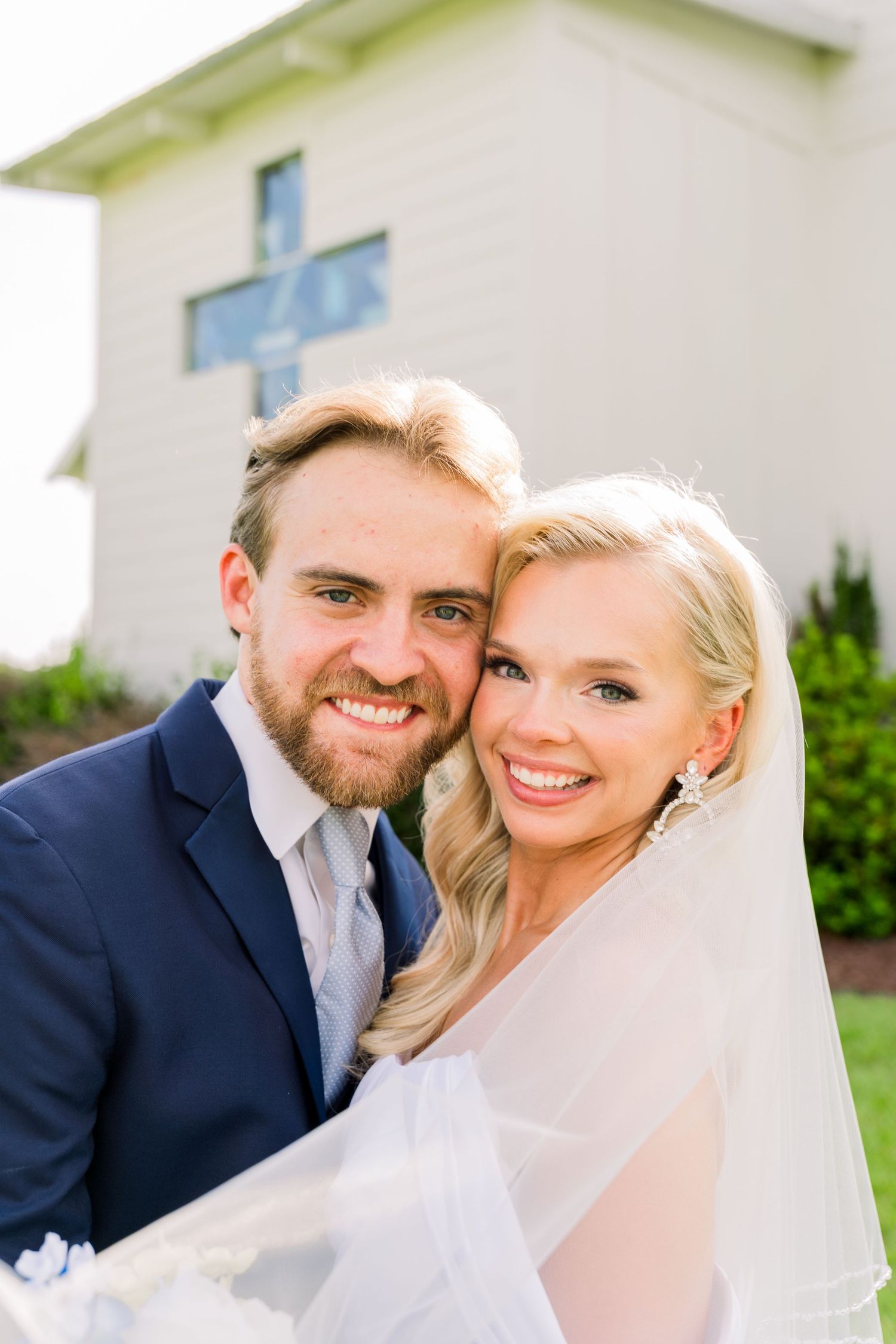 Smiling bride and groom pose cheek-to-cheek outside a white church with a cross, bride wearing a flowing veil.
