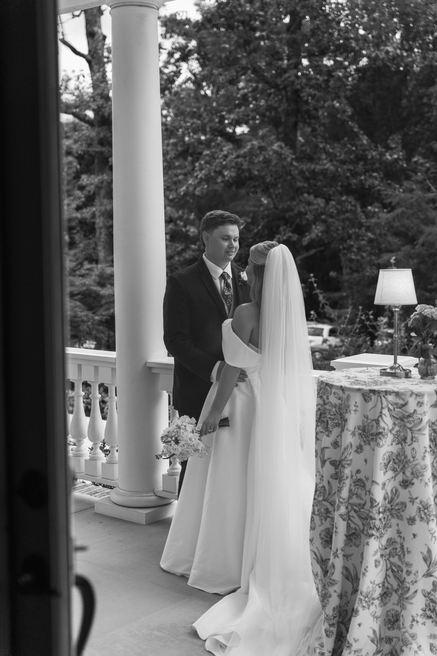 Black and white photo of bride and groom sharing a romantic moment on an elegant outdoor porch with white columns.