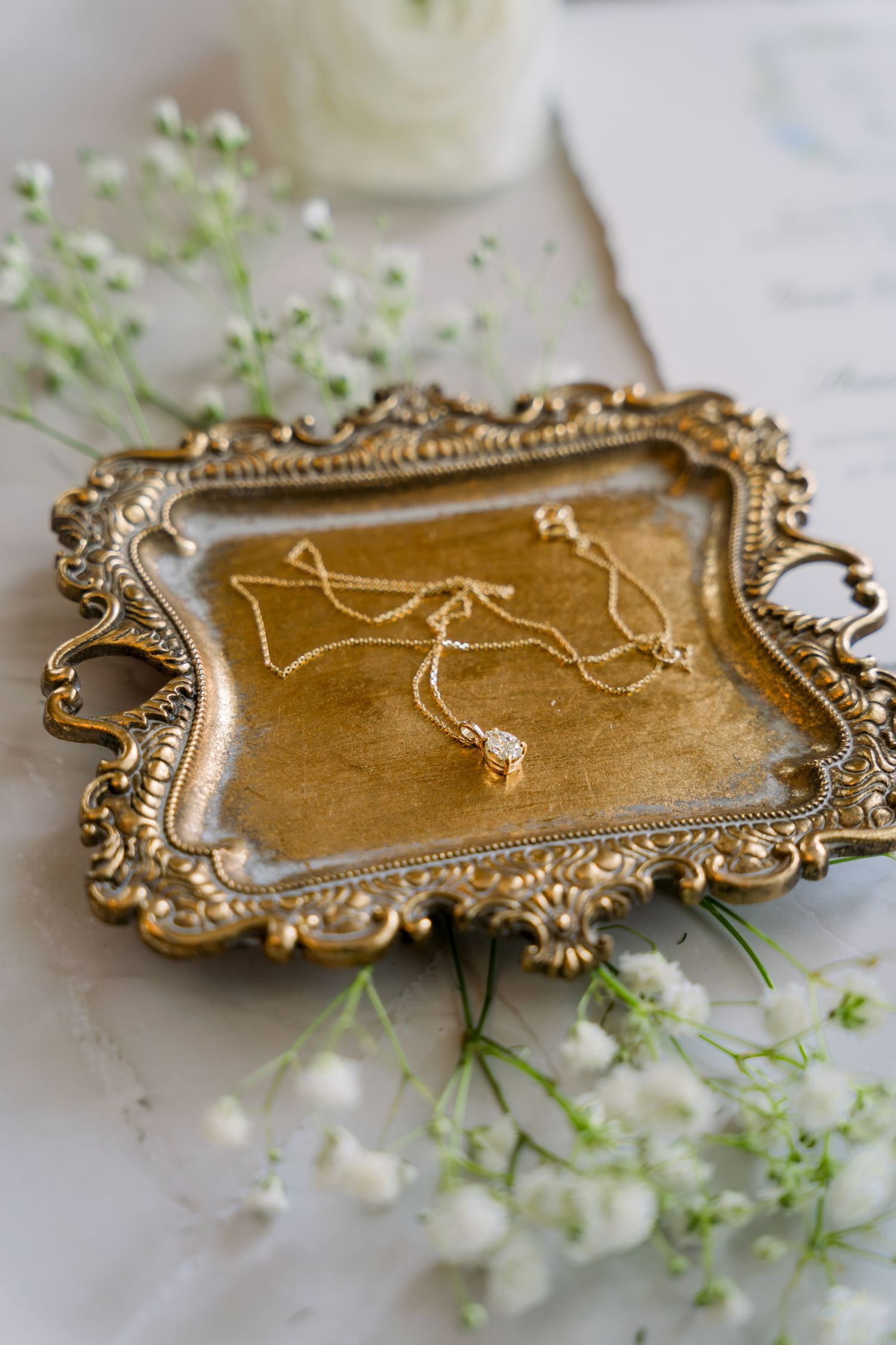 Gold ornate tray with delicate diamond pendant necklace surrounded by white baby's breath flowers on marble surface.
