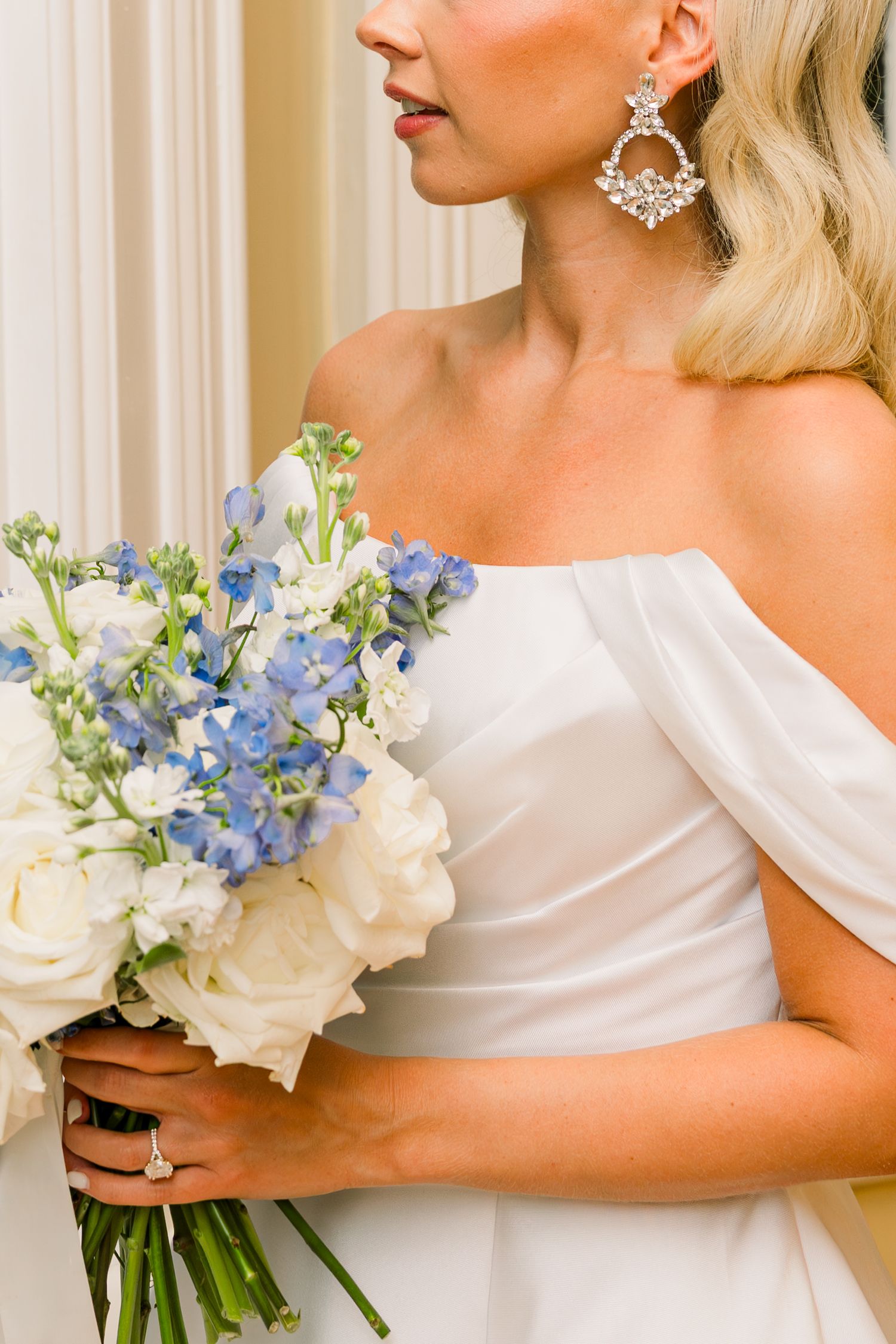 Bride in off-shoulder white gown holding bouquet of white roses and blue flowers, wearing crystal chandelier earrings.