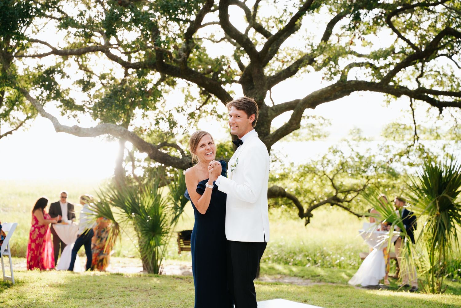 Outdoor wedding reception with groom and mother of the groom dancing under natural tree canopy.