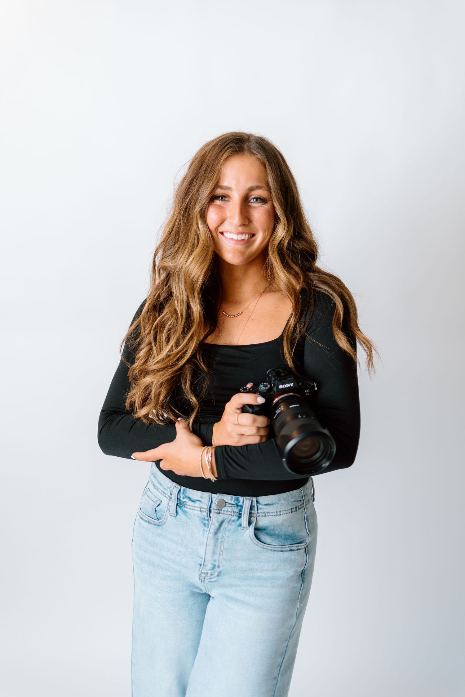 Smiling young woman with wavy brown hair holding a Sony camera, wearing black top and light jeans against white background.