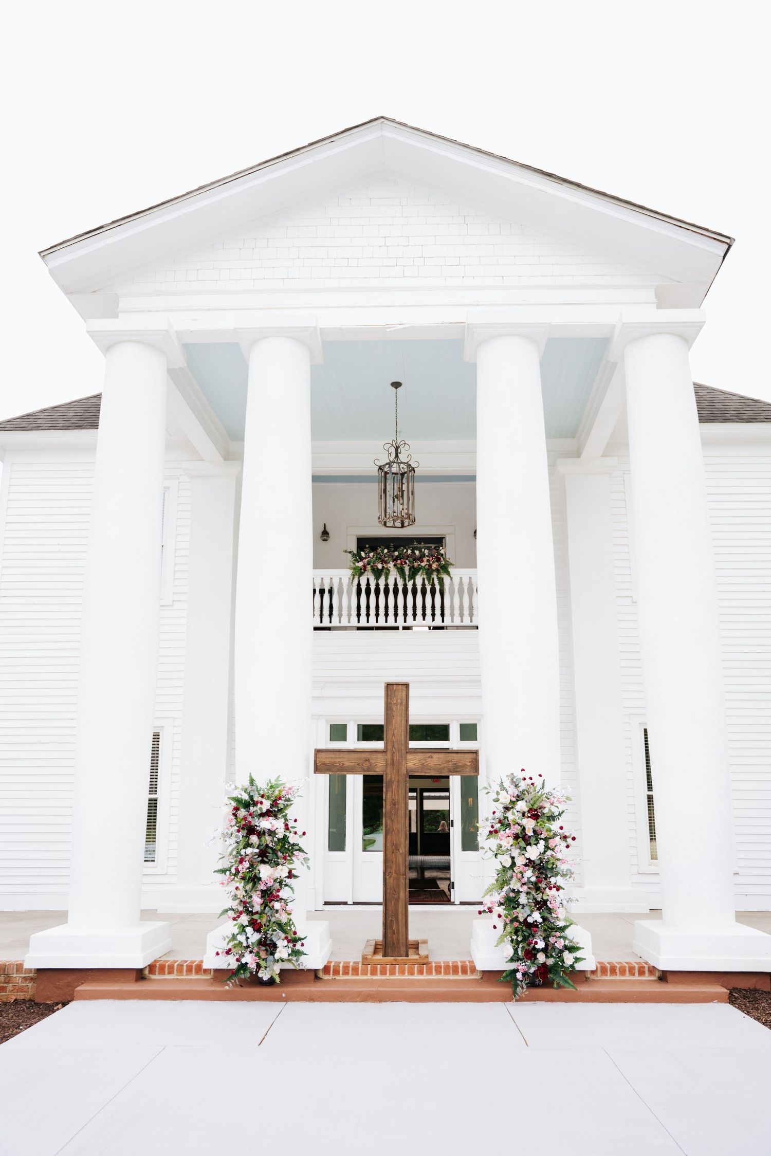 White Southern colonial wedding venue with large columns, wooden cross, and floral arrangements flanking the entrance.