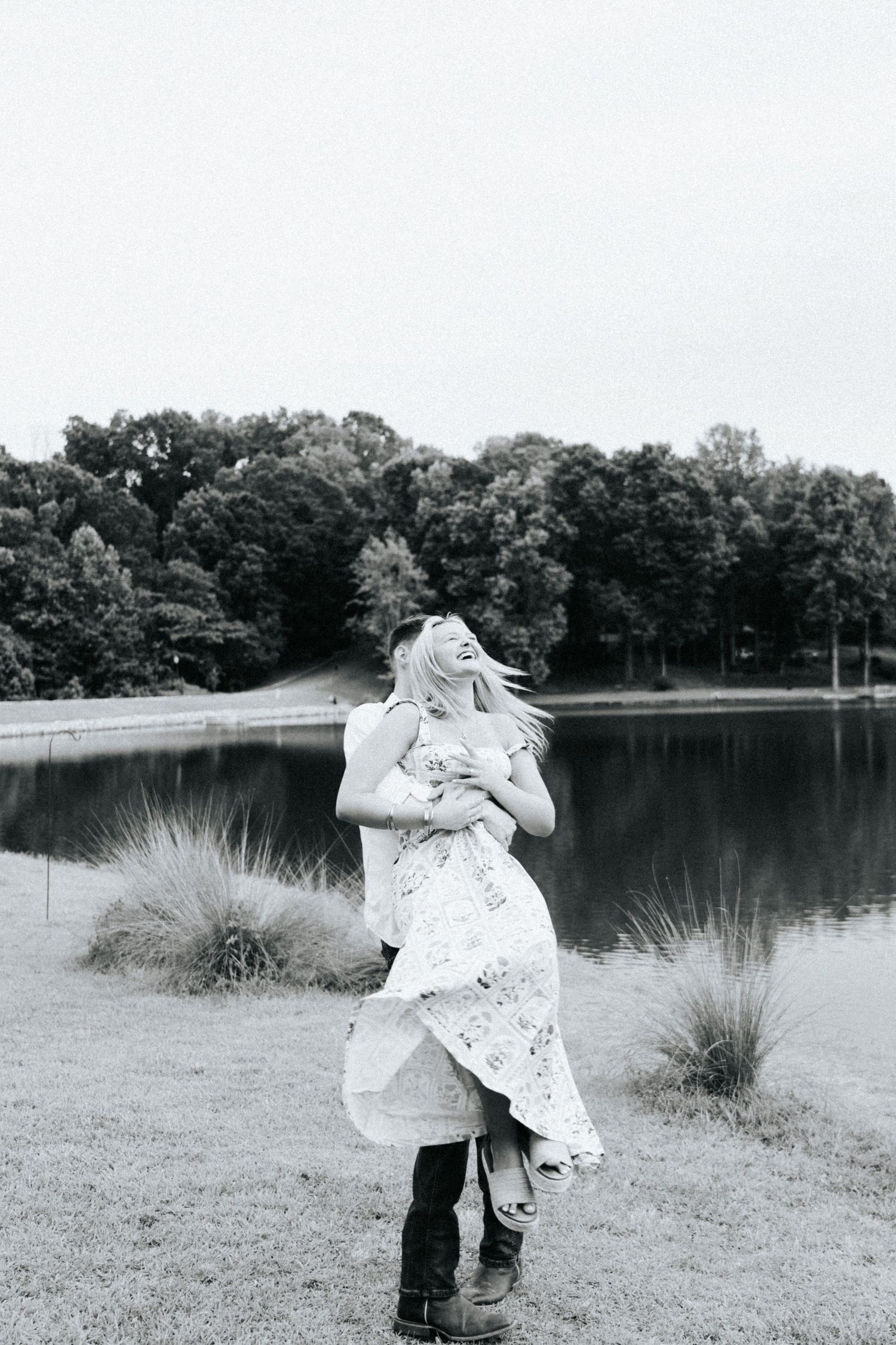 Black and white photo of a couple embracing by a tranquil lake with tall grass.