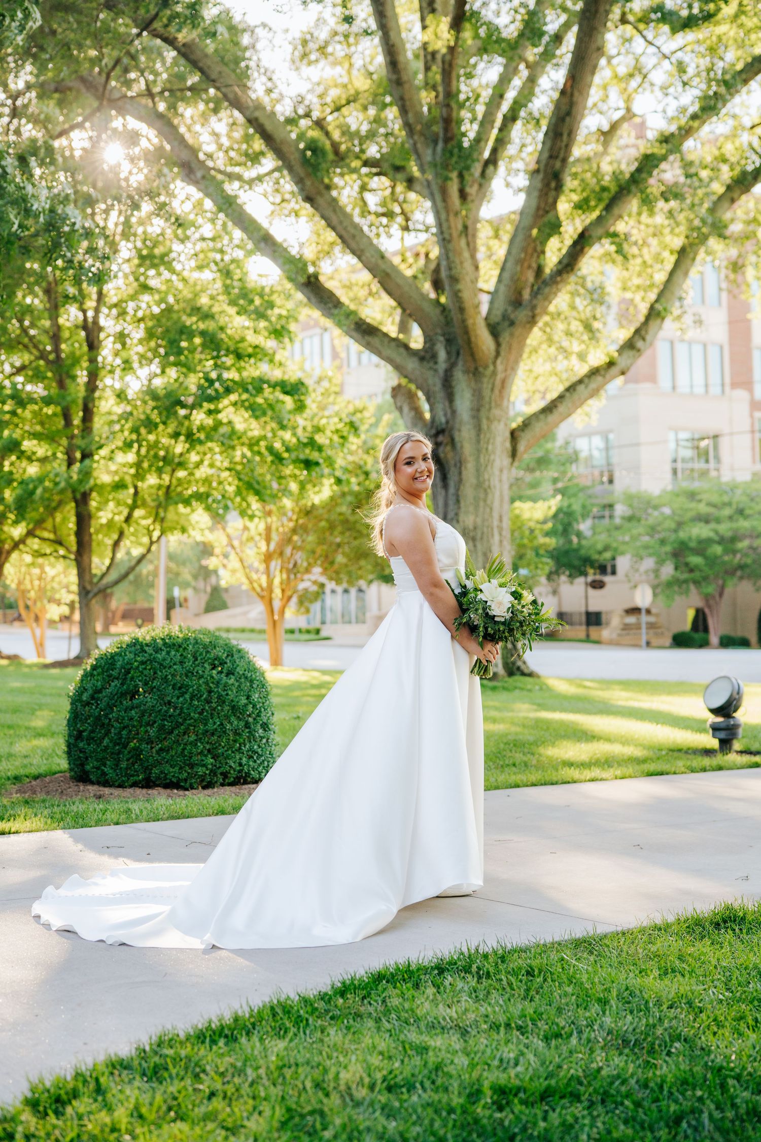 Sunlit bridal portrait on church grounds with flowing white dress and mature trees in background.