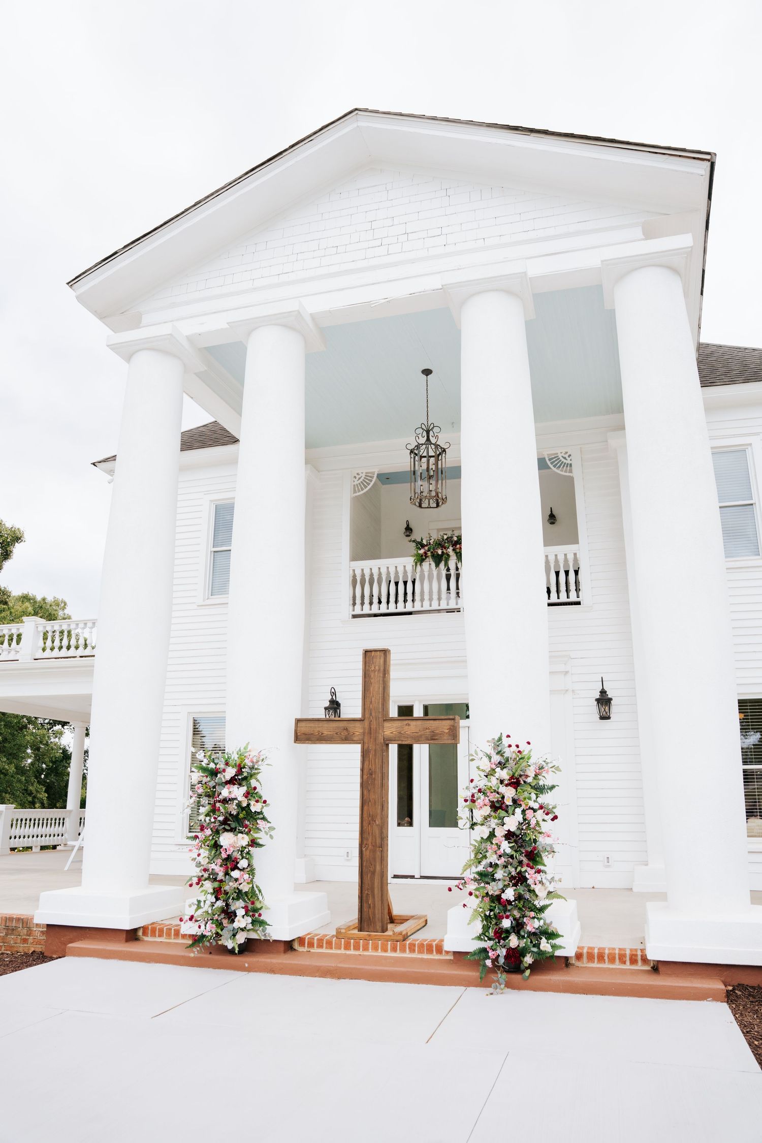 Elegant white church entrance with tall columns, wooden cross, and pink floral arrangements on brick steps.