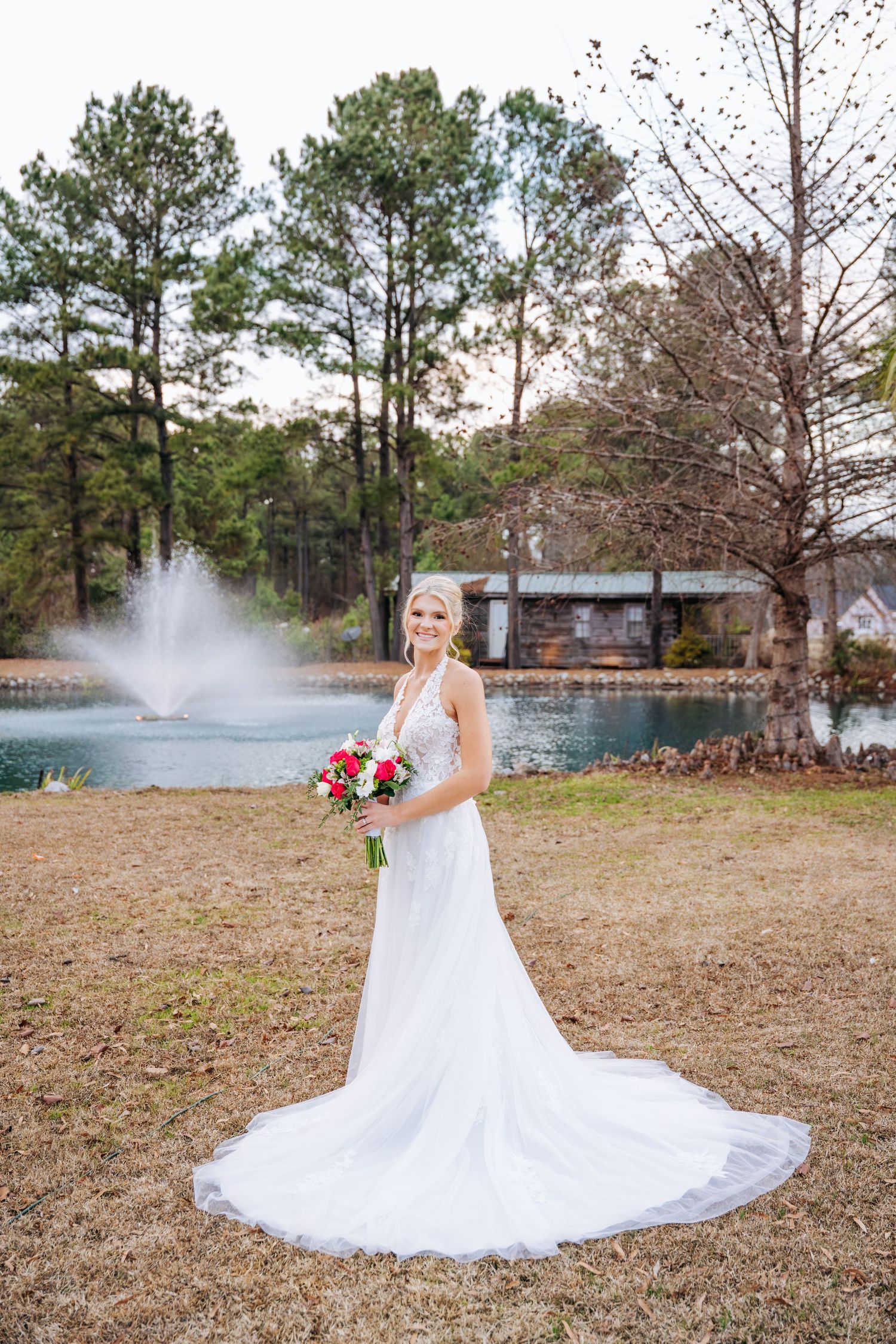 A bride in a flowing white wedding dress poses gracefully in front of a misty lake and pine trees on a fall day.