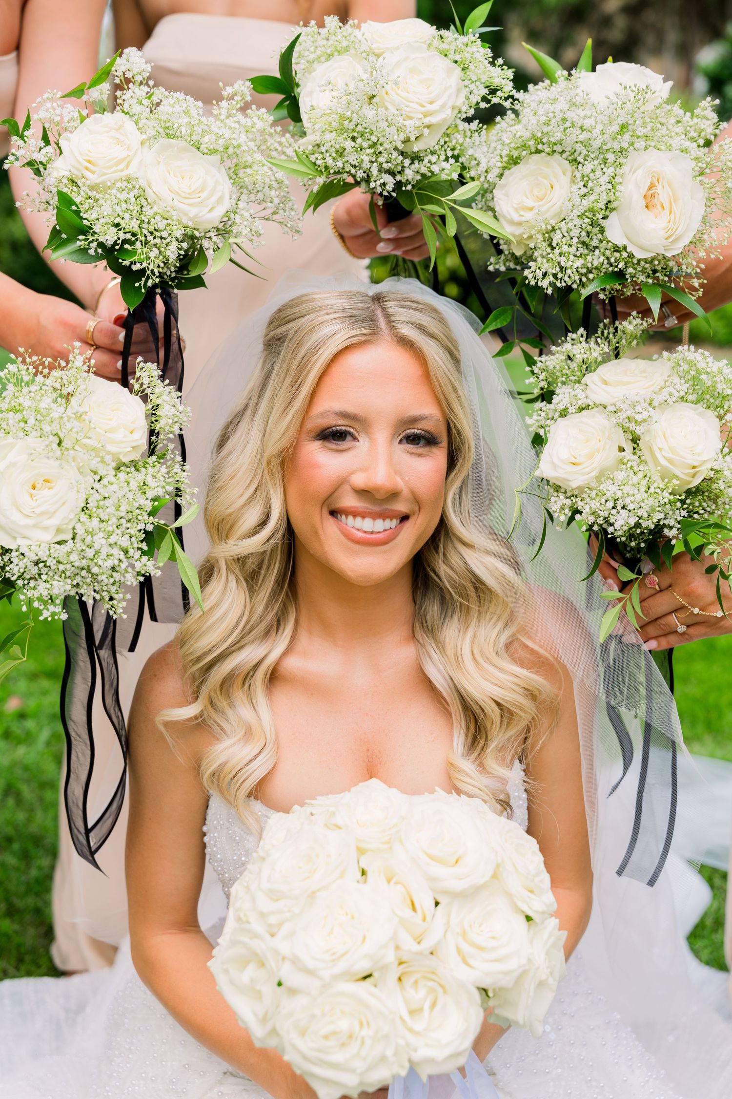 Smiling bride holding white rose bouquet surrounded by bridesmaids with matching white rose and baby's breath bouquets.