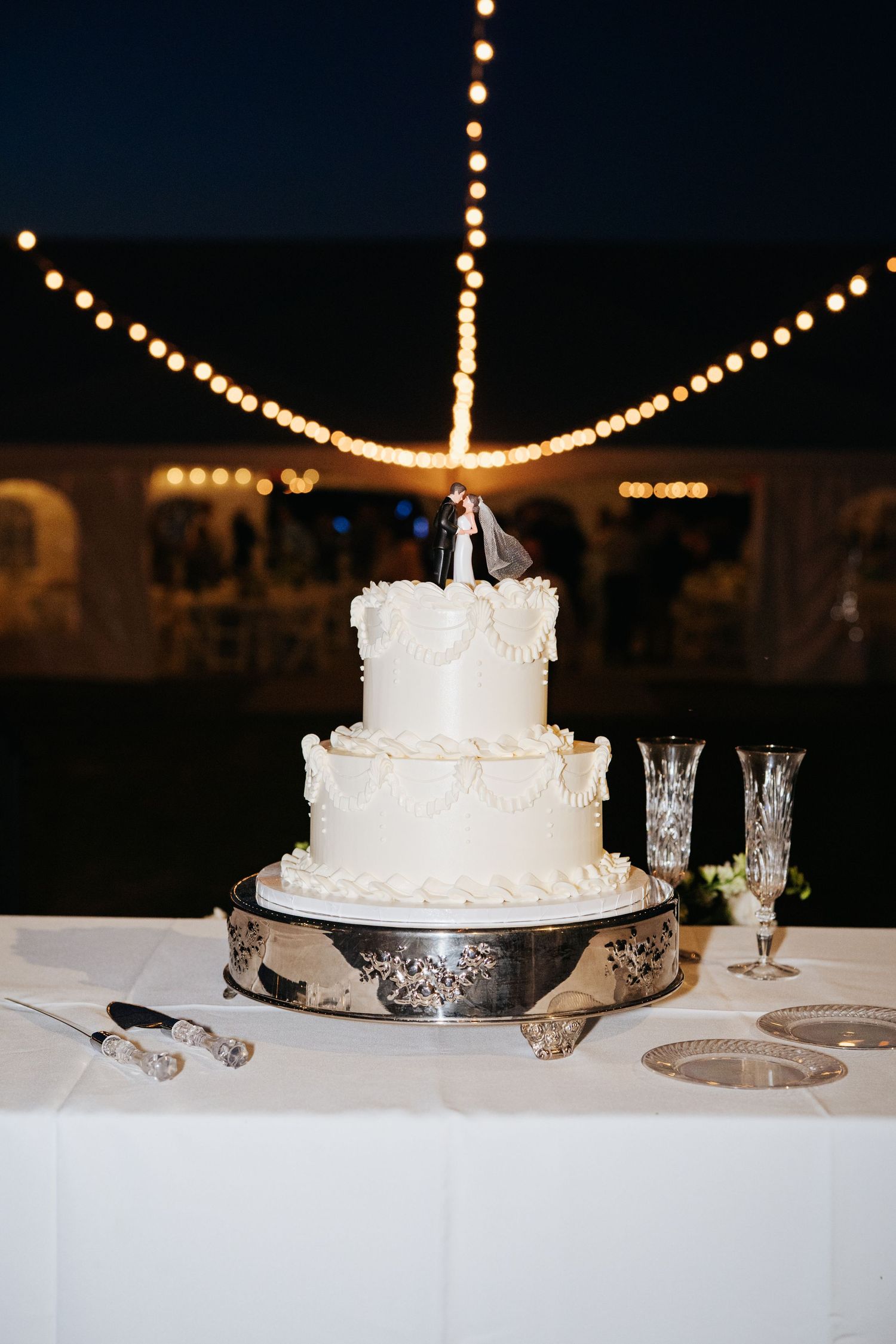White wedding cake on display table with string lights glowing in the background.