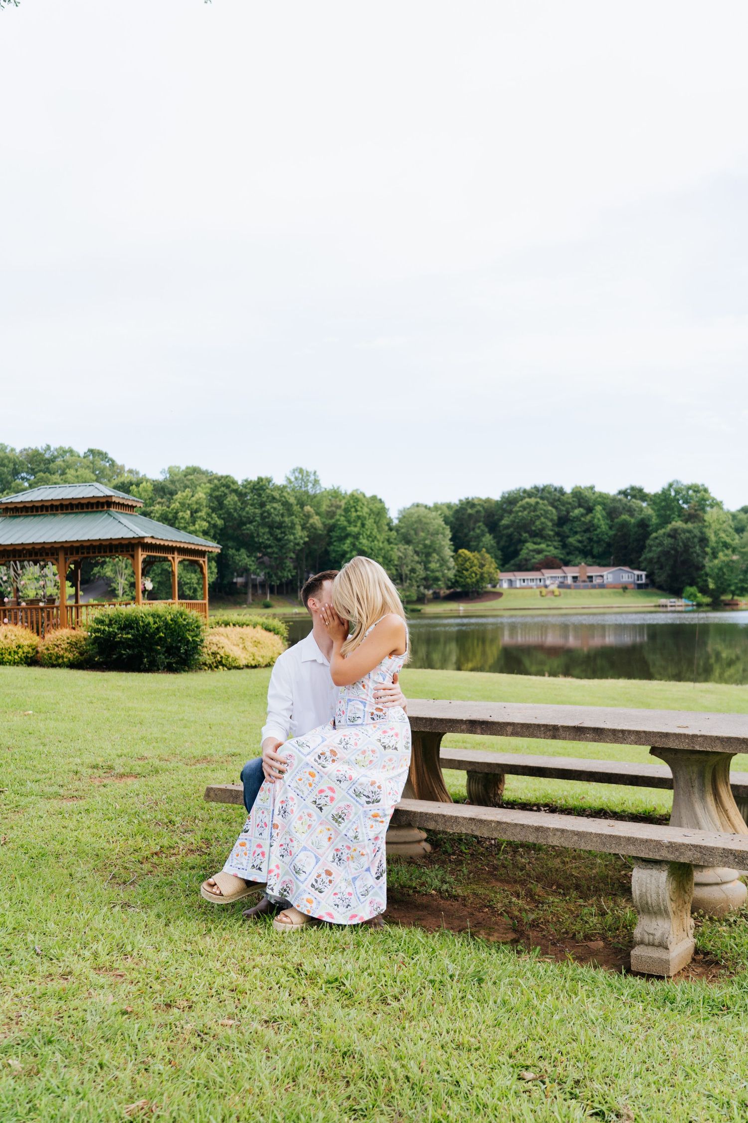 A floral dress catches the breeze as someone sits on a stone bench near a lakeside gazebo.