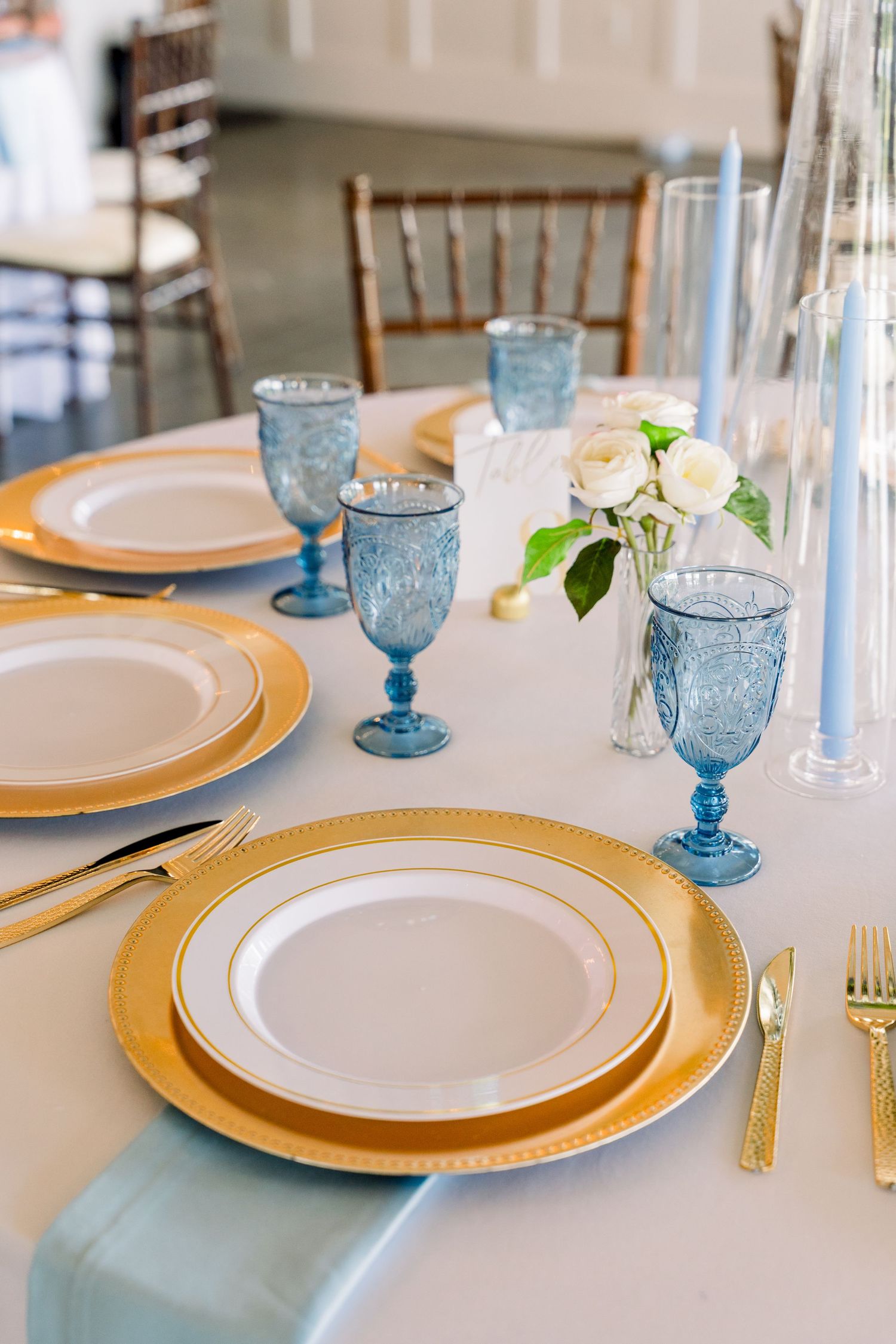 Elegant wedding table setting with gold-rimmed plates, blue goblets, gold cutlery, and white roses on a light blue tablecloth.