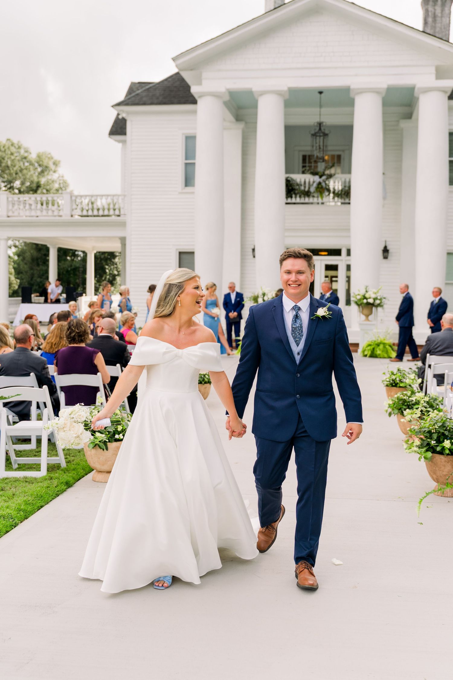 Bride in off-shoulder gown and groom in navy suit walk hand-in-hand down aisle at elegant white mansion wedding.