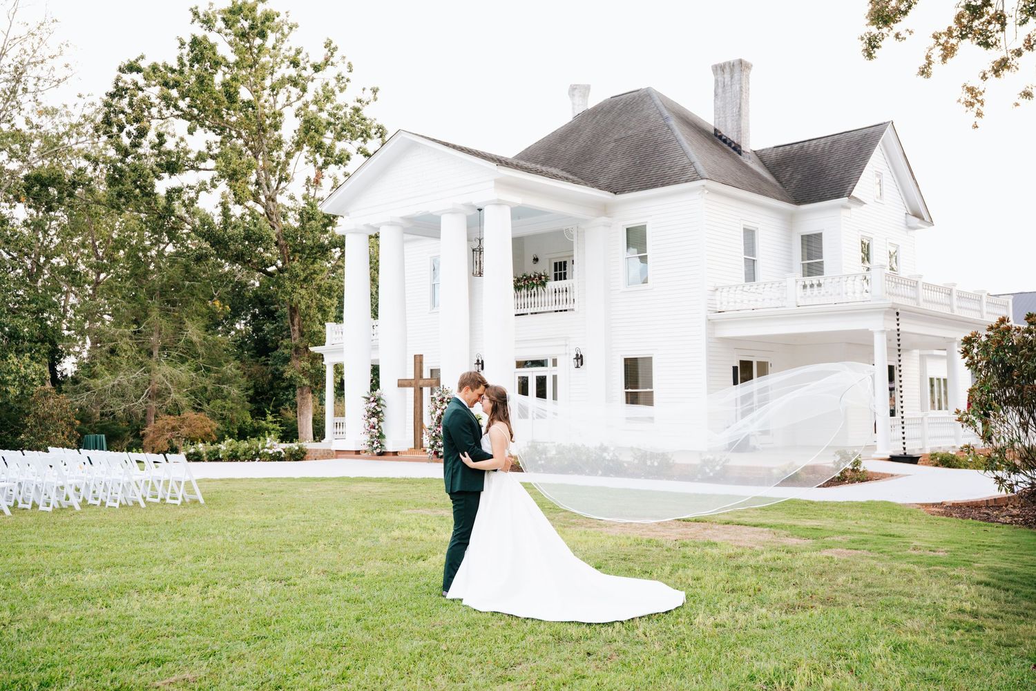 A flowing white wedding dress billows in the wind on a lush green lawn in front of a classic white farmhouse.