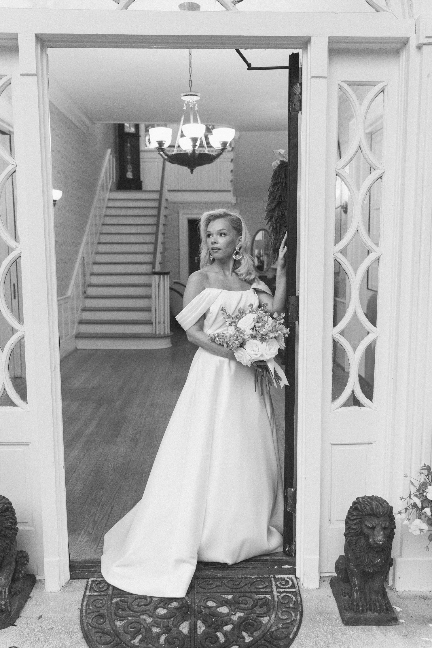 Bride in off-shoulder white gown holds bouquet at elegant home entrance with chandelier and staircase in black and white.