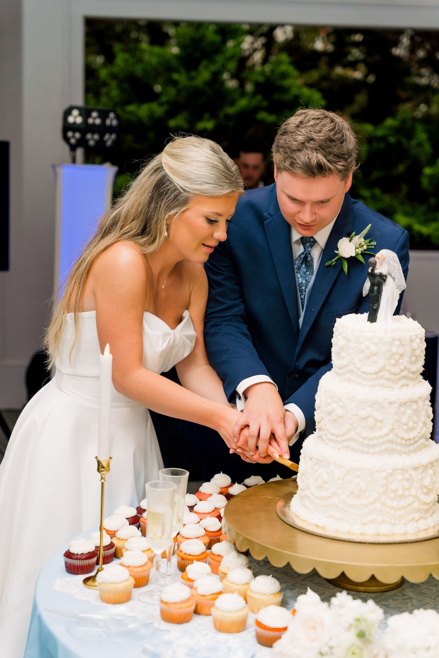 Bride and groom cutting a white three-tier wedding cake together at outdoor reception with cupcakes nearby.