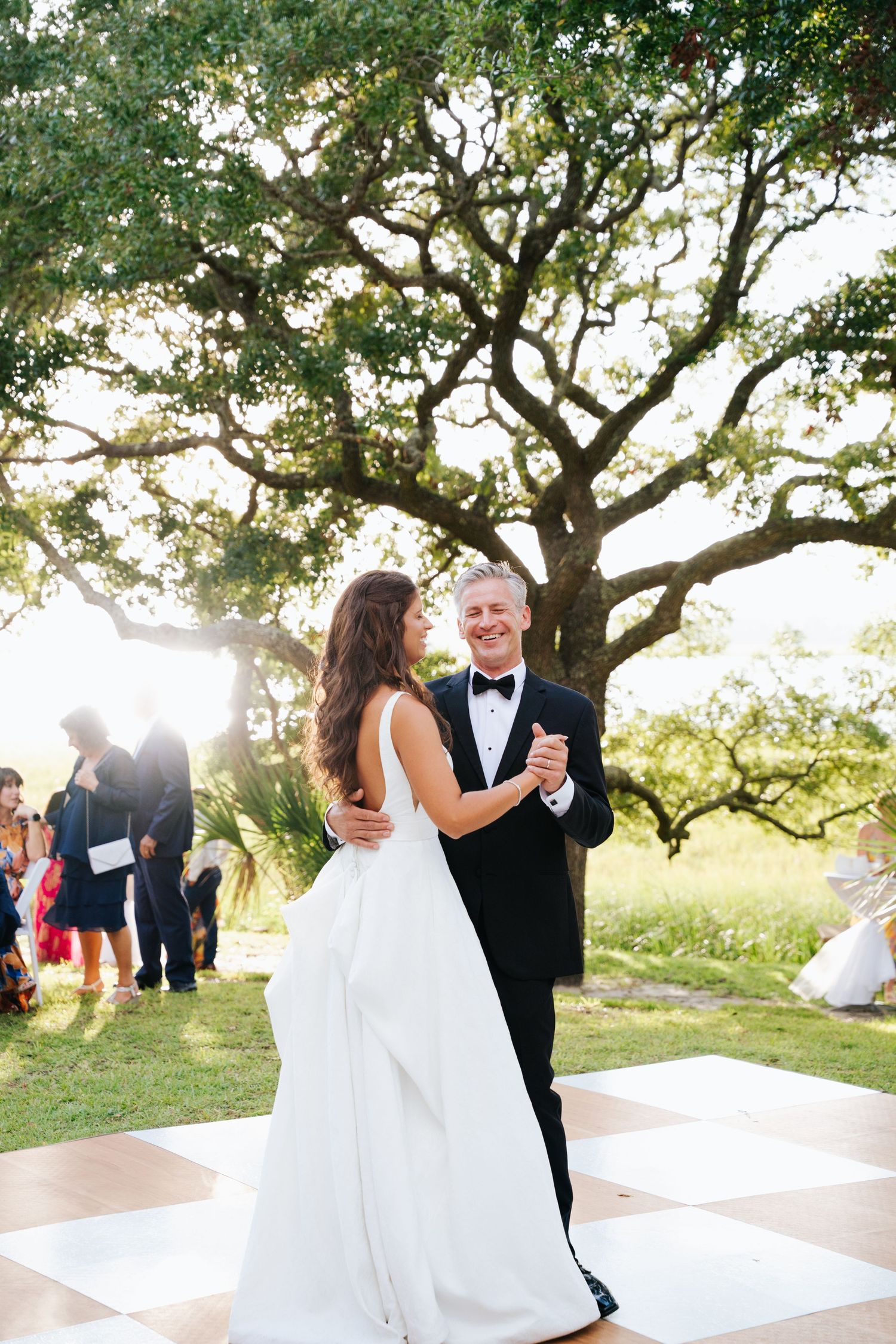 Bride and father are dancing on an outdoor checkered dance floor beneath sprawling oak trees.