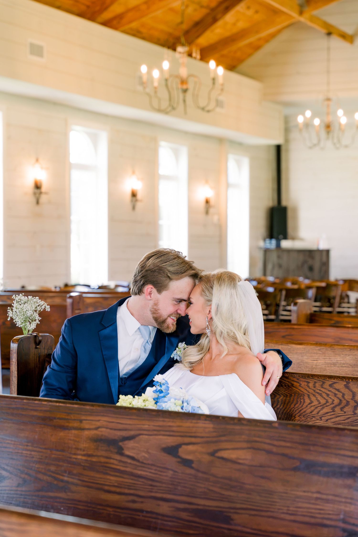 Bride and groom sharing an intimate moment on wooden church pews, groom in navy suit, bride holding blue and white bouquet.