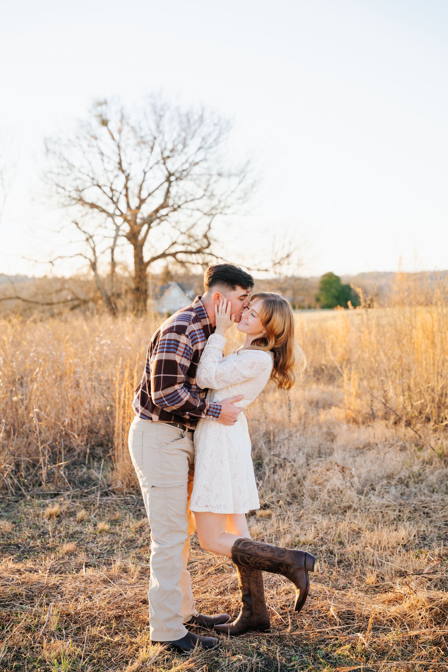 Two people share a tender moment in a golden field at sunset with bare trees in the background.