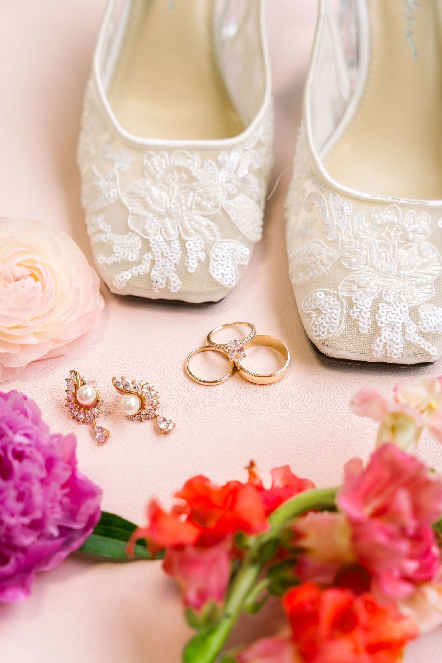 Elegant bridal flat shoes with lace, gold wedding rings, pearl earrings, and colorful flowers on pink background.
