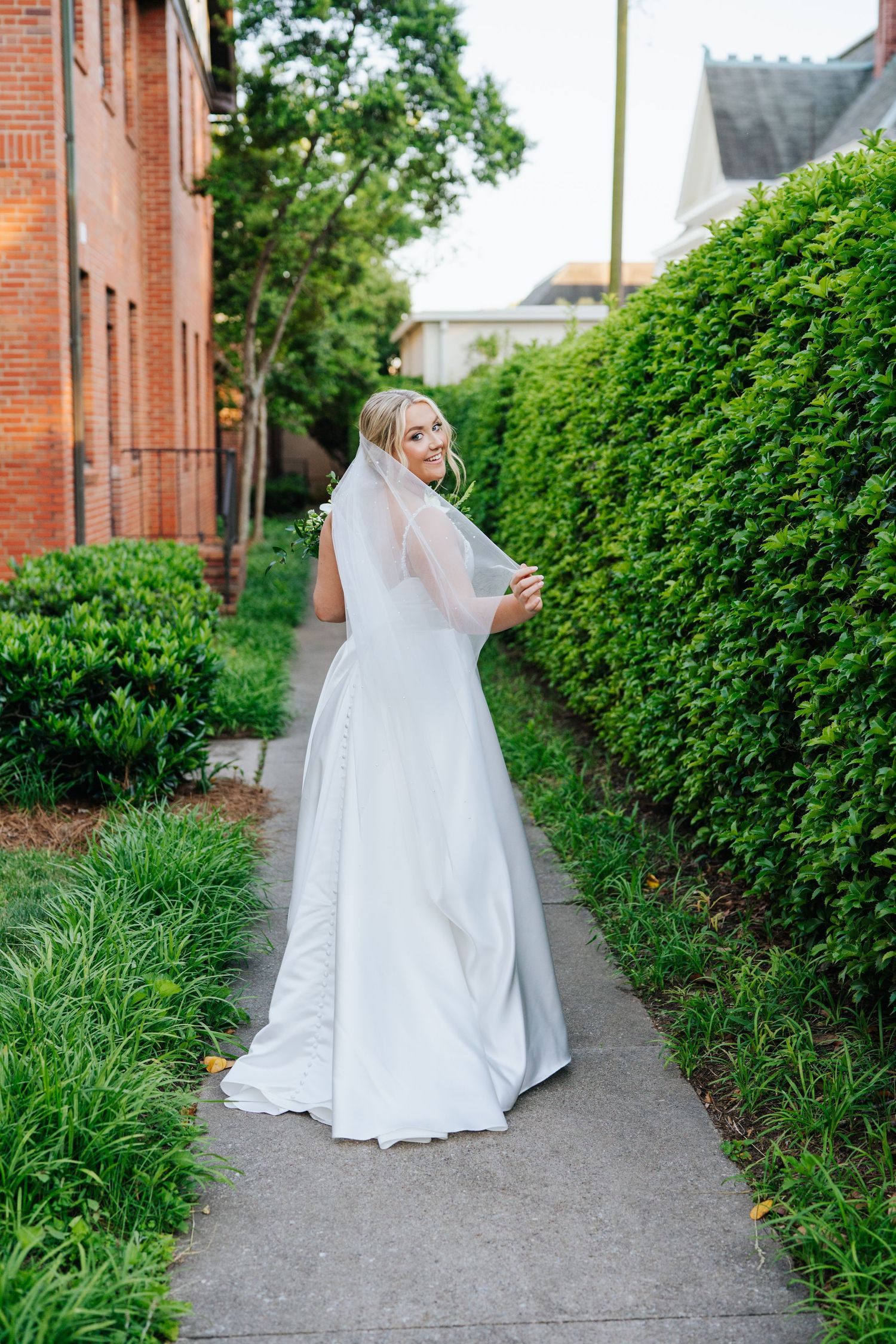 A bride poses on a garden path between brick buildings and manicured hedges.