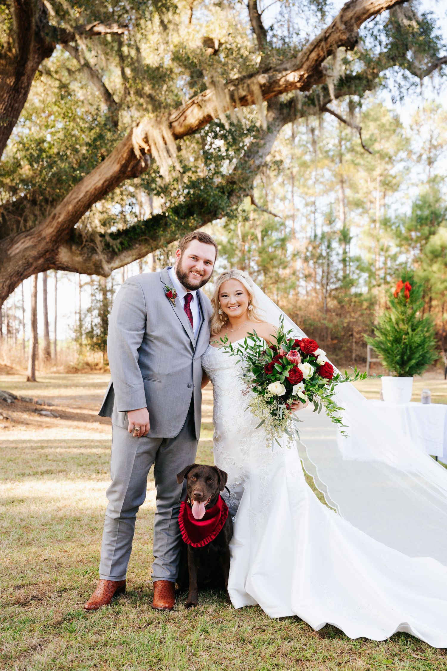 Bride and groom smile outdoors under a mossy oak tree, holding red bouquet, with their chocolate Lab wearing a red lei.